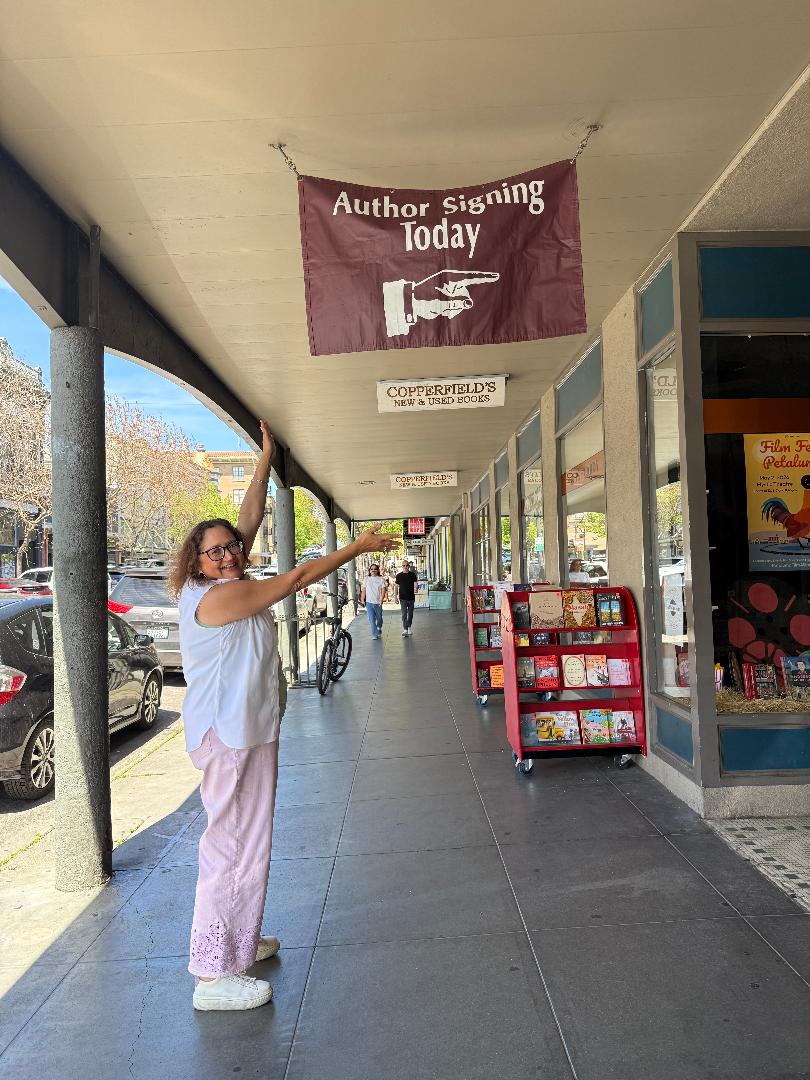 Woman pointing at a sign that says 'Author Signing Today' on a sidewalk outside a bookstore.