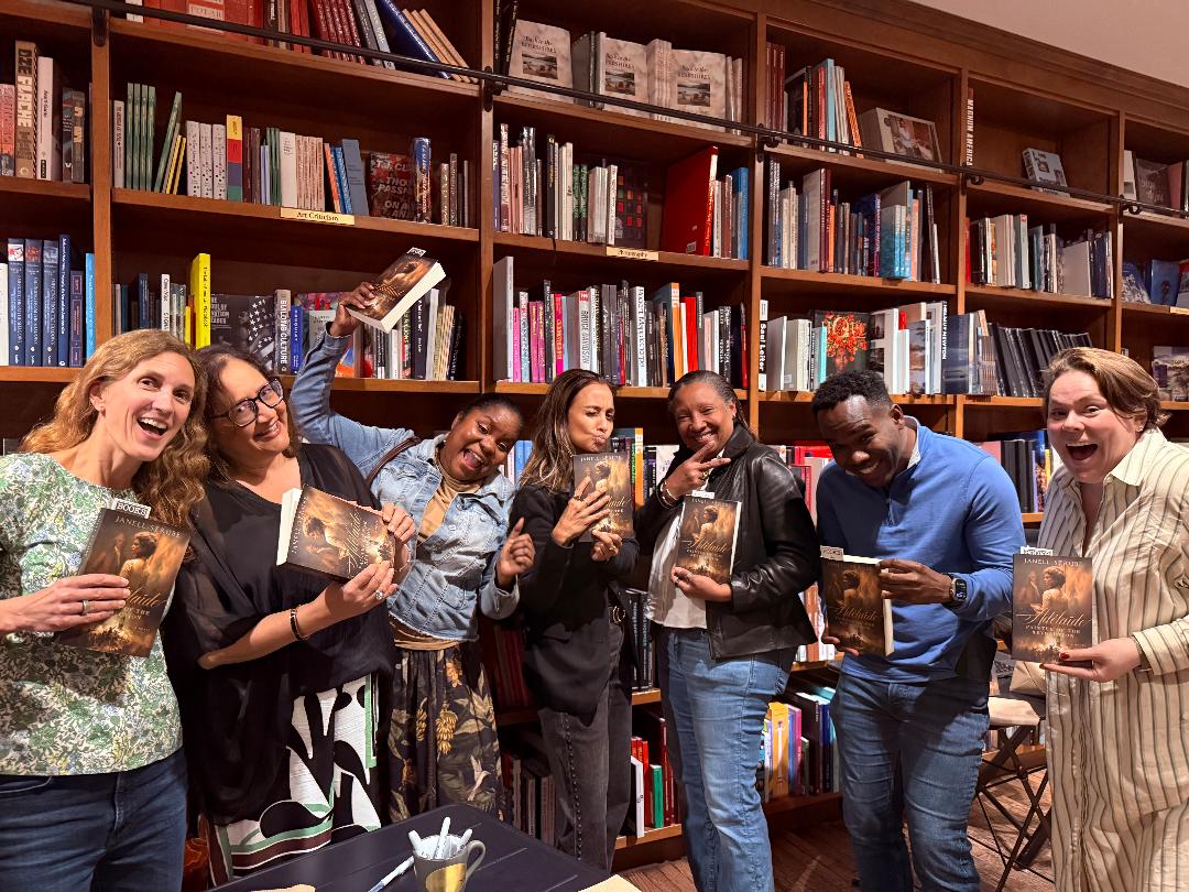 Group of seven people in a library, holding books and smiling, with shelves filled with books in the background.