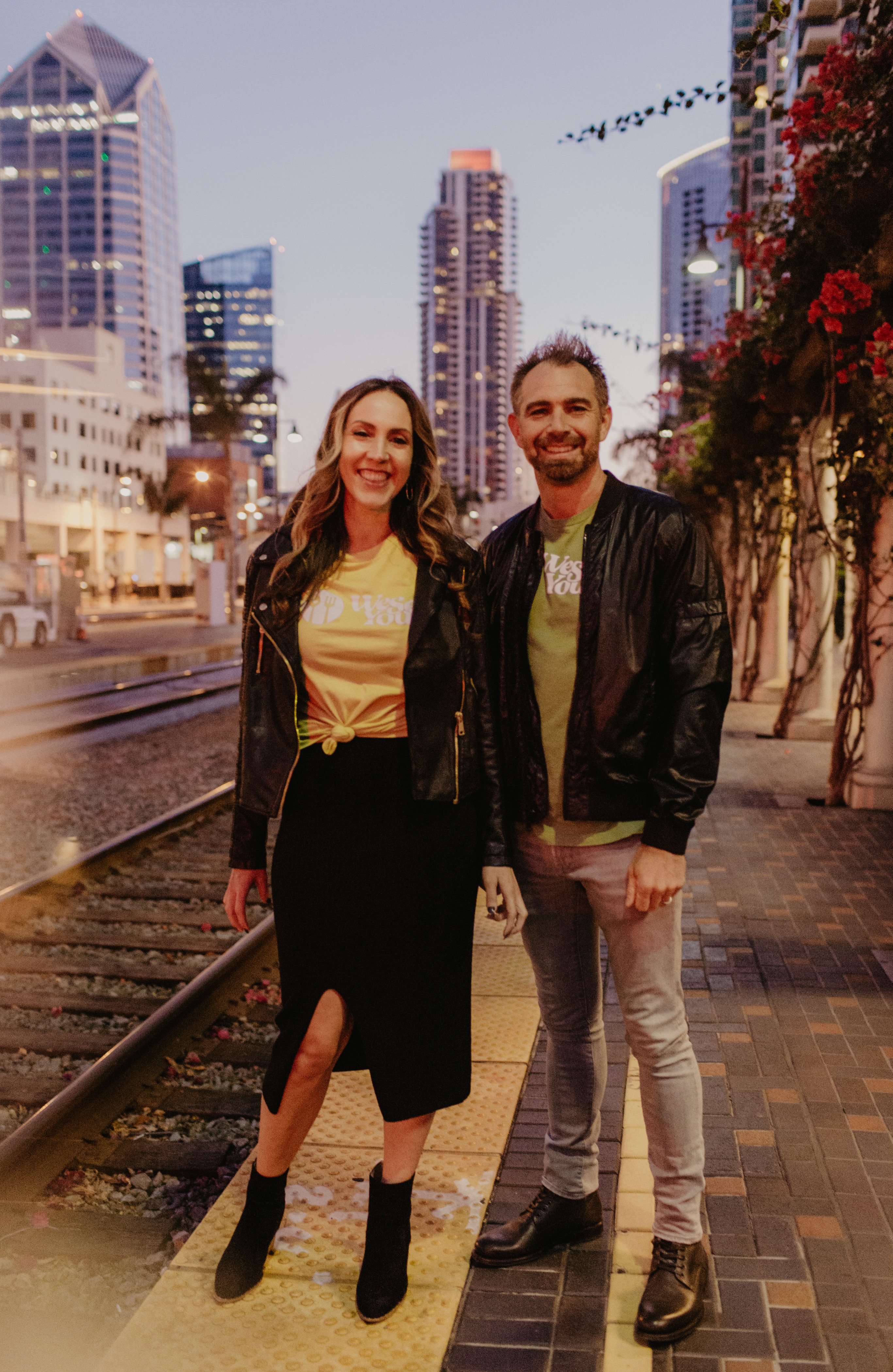 A smiling woman and man stand on a city sidewalk at dusk, with tall buildings and flowers in the background.