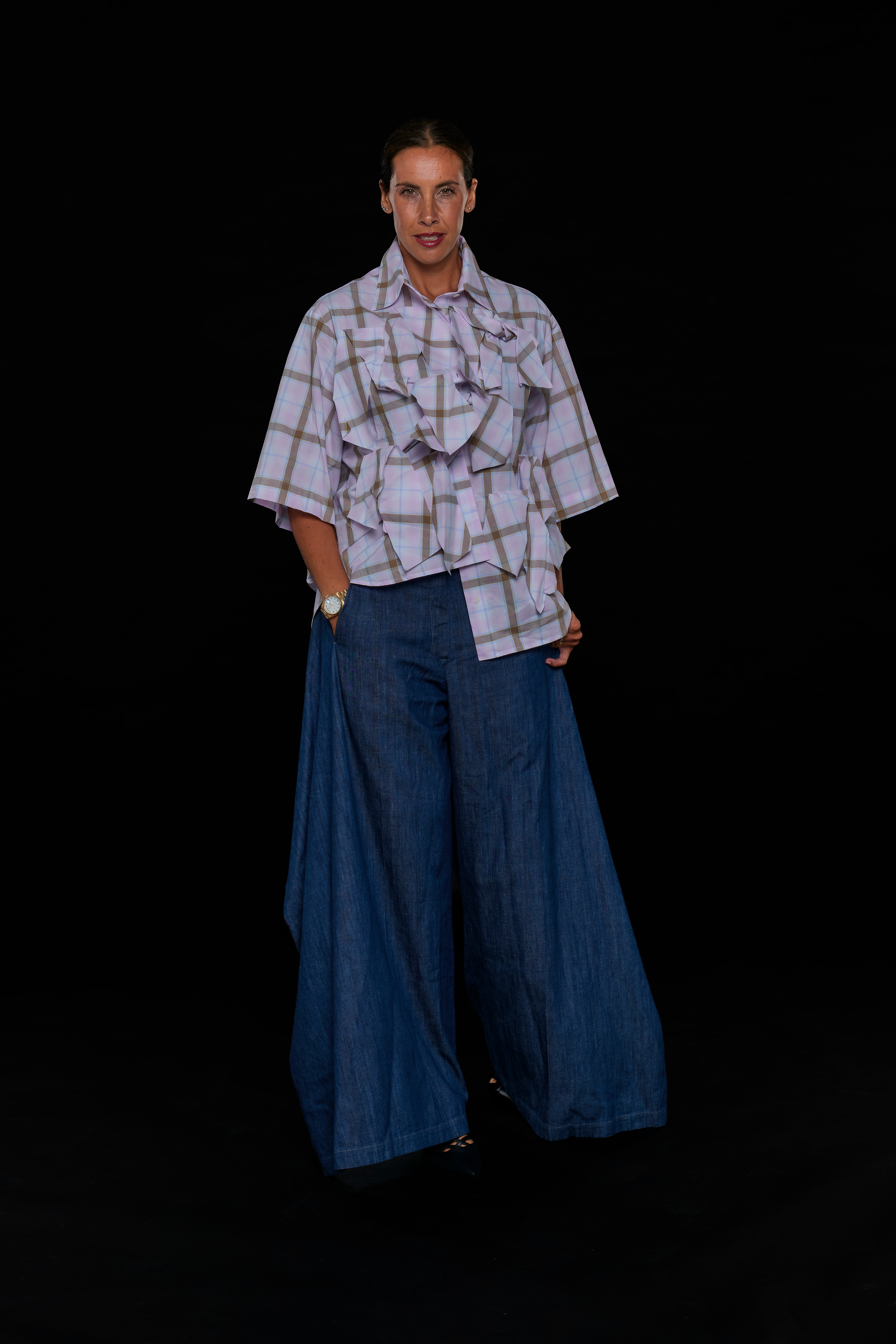 Woman standing against black background, wearing a checkered shirt and wide-leg jeans, with hands in pockets.