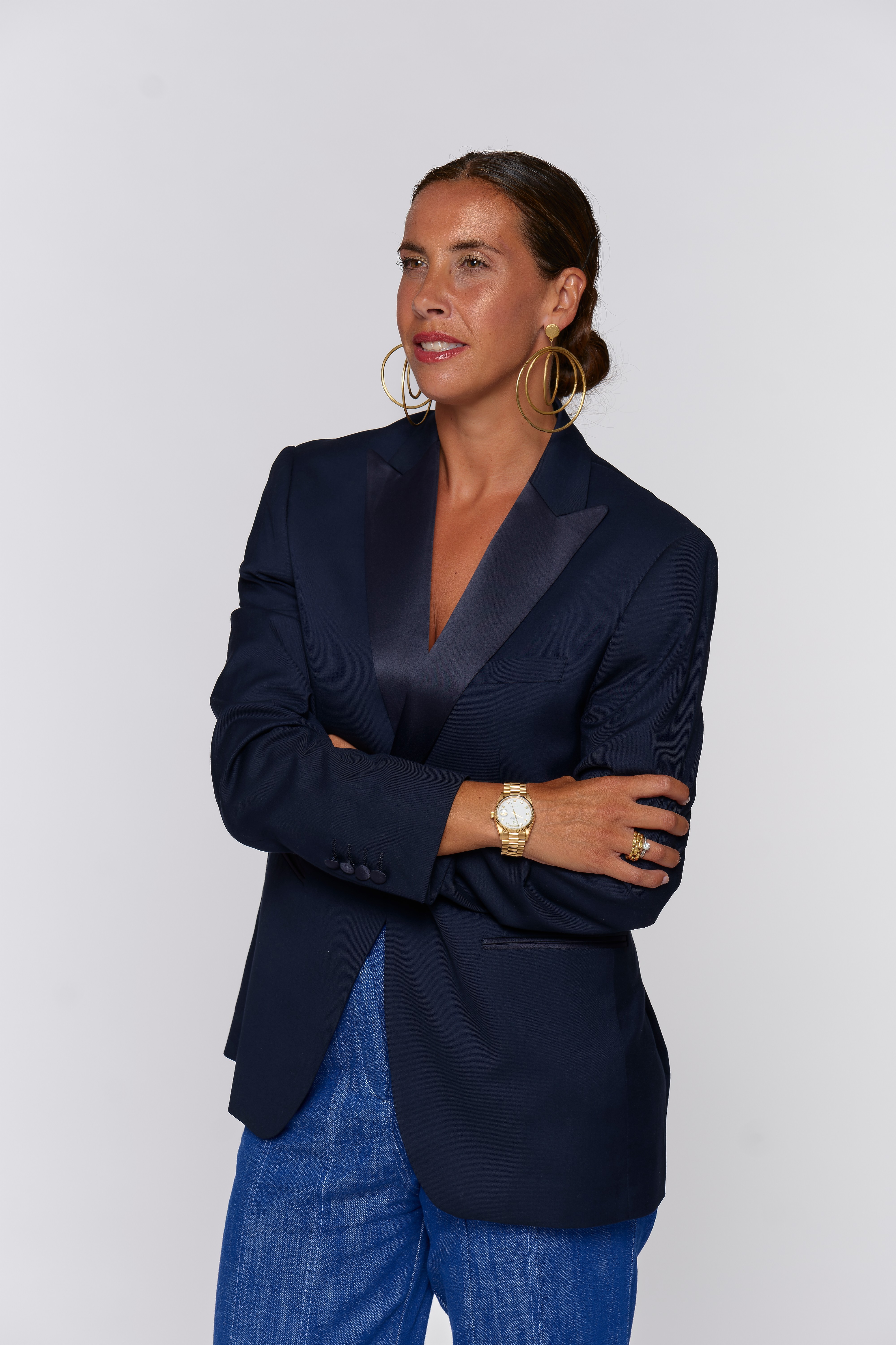 Woman with dark hair in a bun wearing a navy blazer, blue jeans, and a watch, standing with arms crossed.