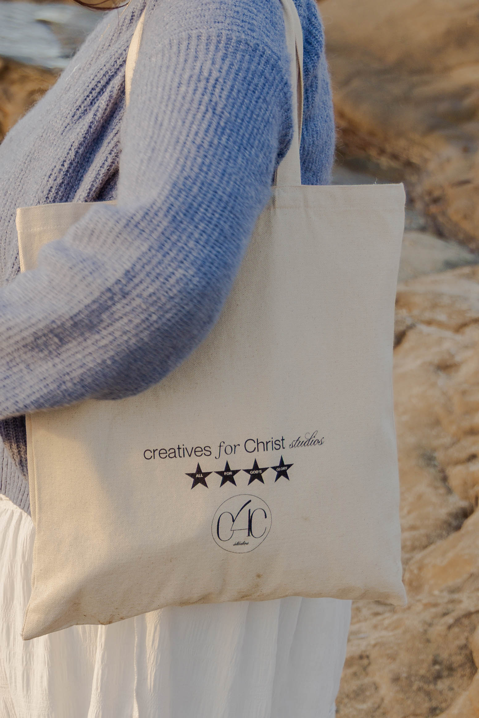 Person wearing a blue sweater holding a white tote bag with text and stars, outdoors with rocks in background.