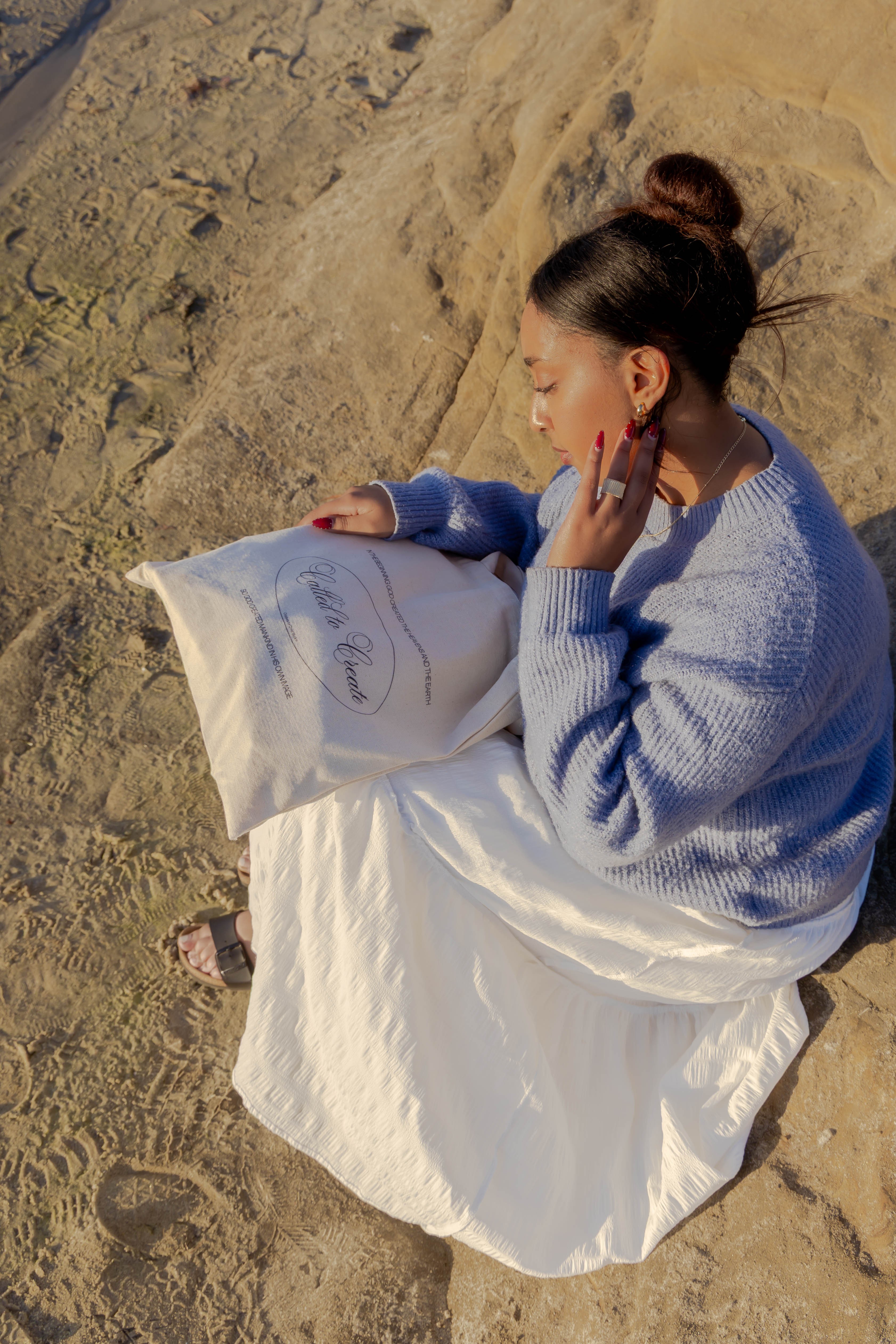 Woman sitting on sand, reading a book, wearing a blue sweater and white skirt, with her hair in a bun.