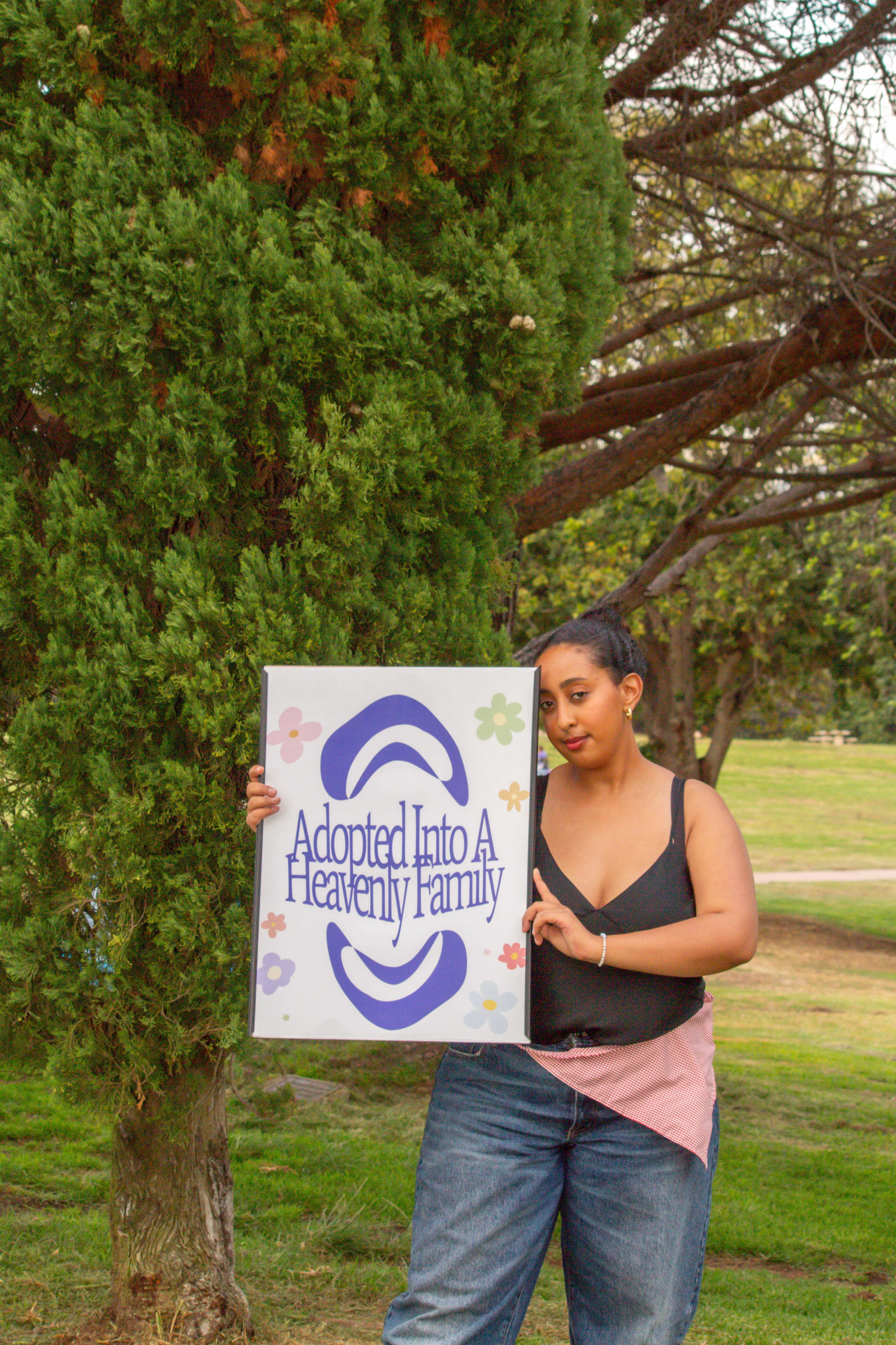 Young woman holding a sign that reads 'Adopted Into A Healthy Family' outdoors with trees and grass.