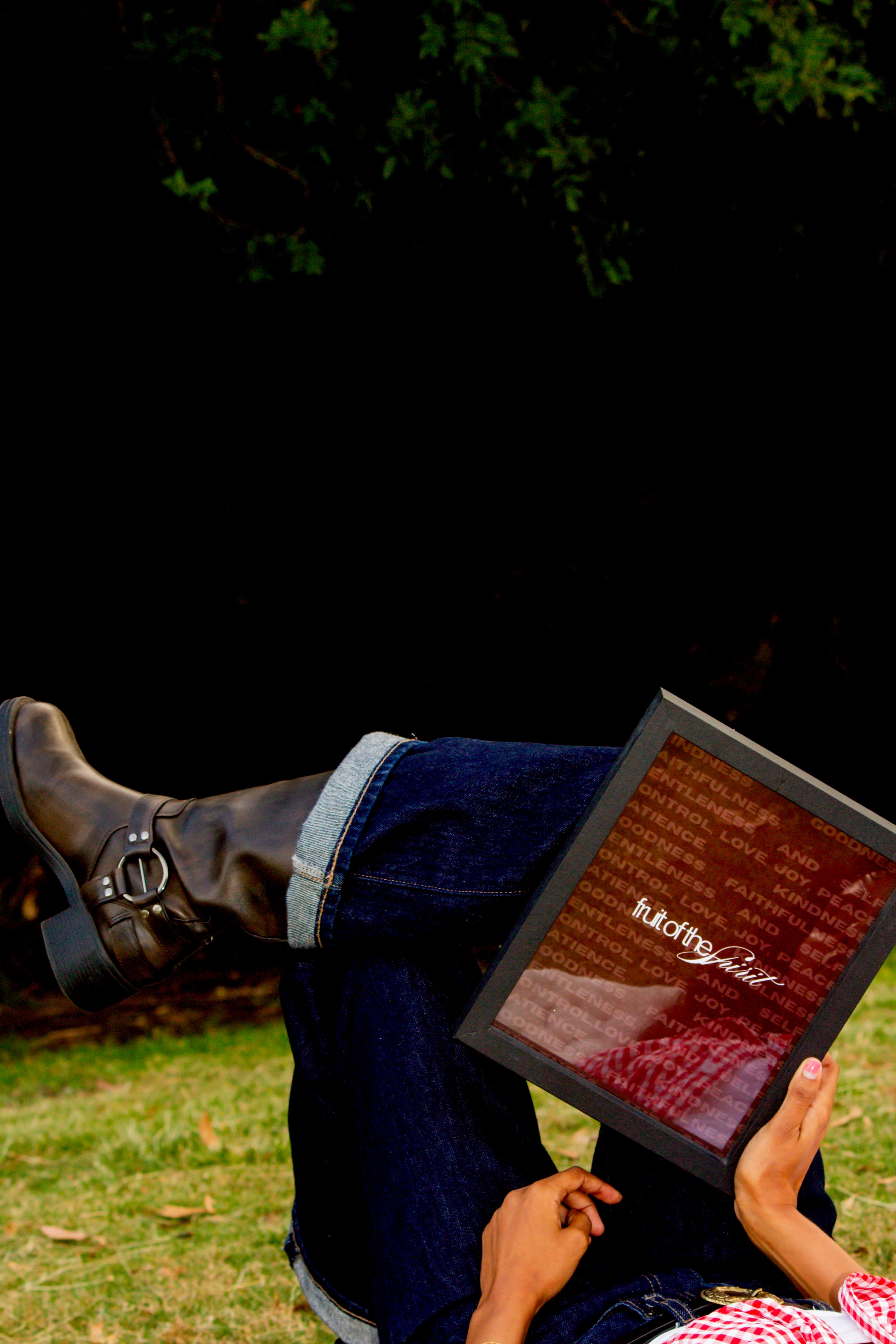 Person sitting outdoors with legs crossed, holding a tablet displaying red text, surrounded by greenery.