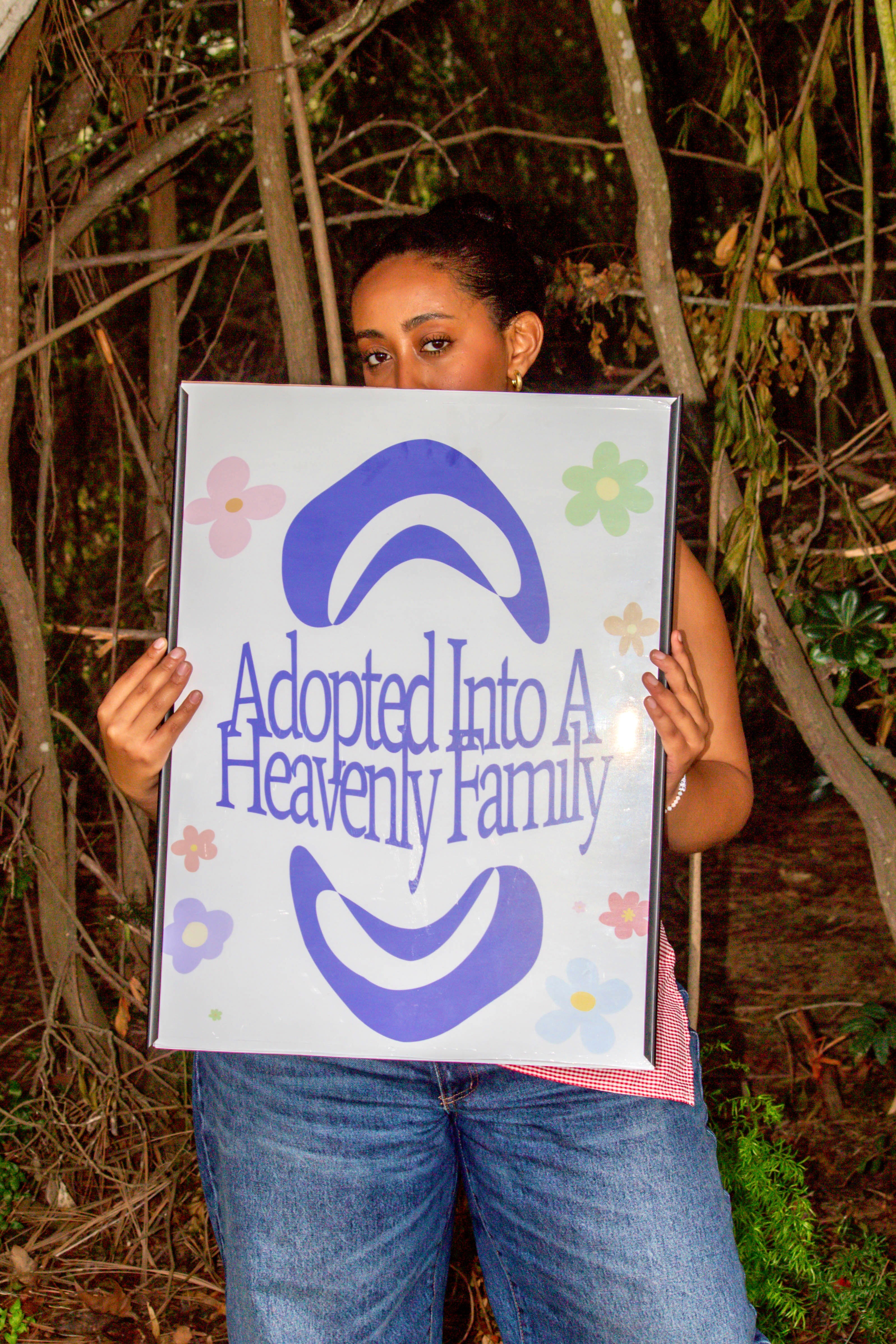 Person holding a sign with text and colorful flowers outdoors, partially obscured by the sign, surrounded by trees and plants.