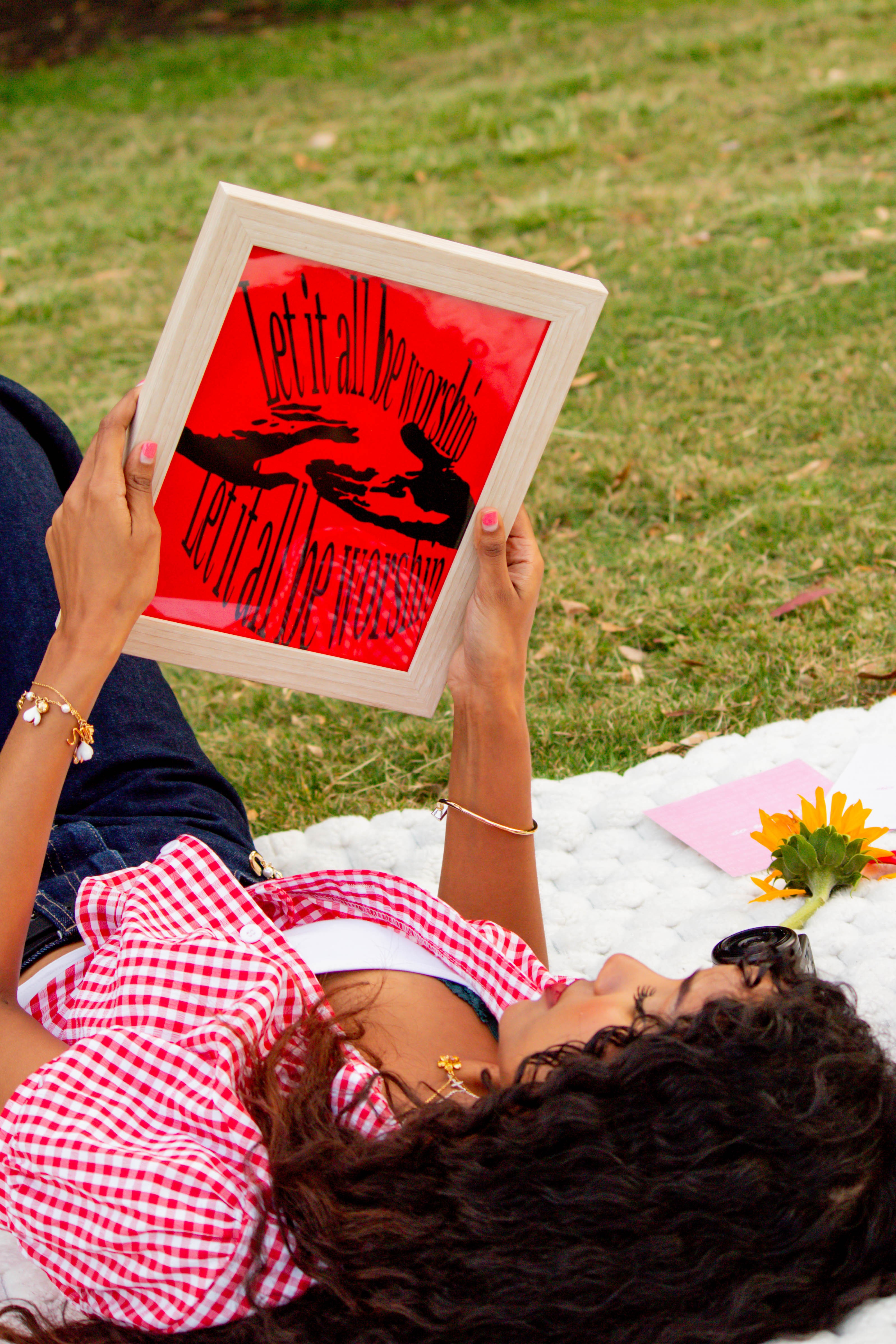 Person lying on grass holding a tablet displaying a red and black graphic with text, surrounded by flowers and a picnic blanket.