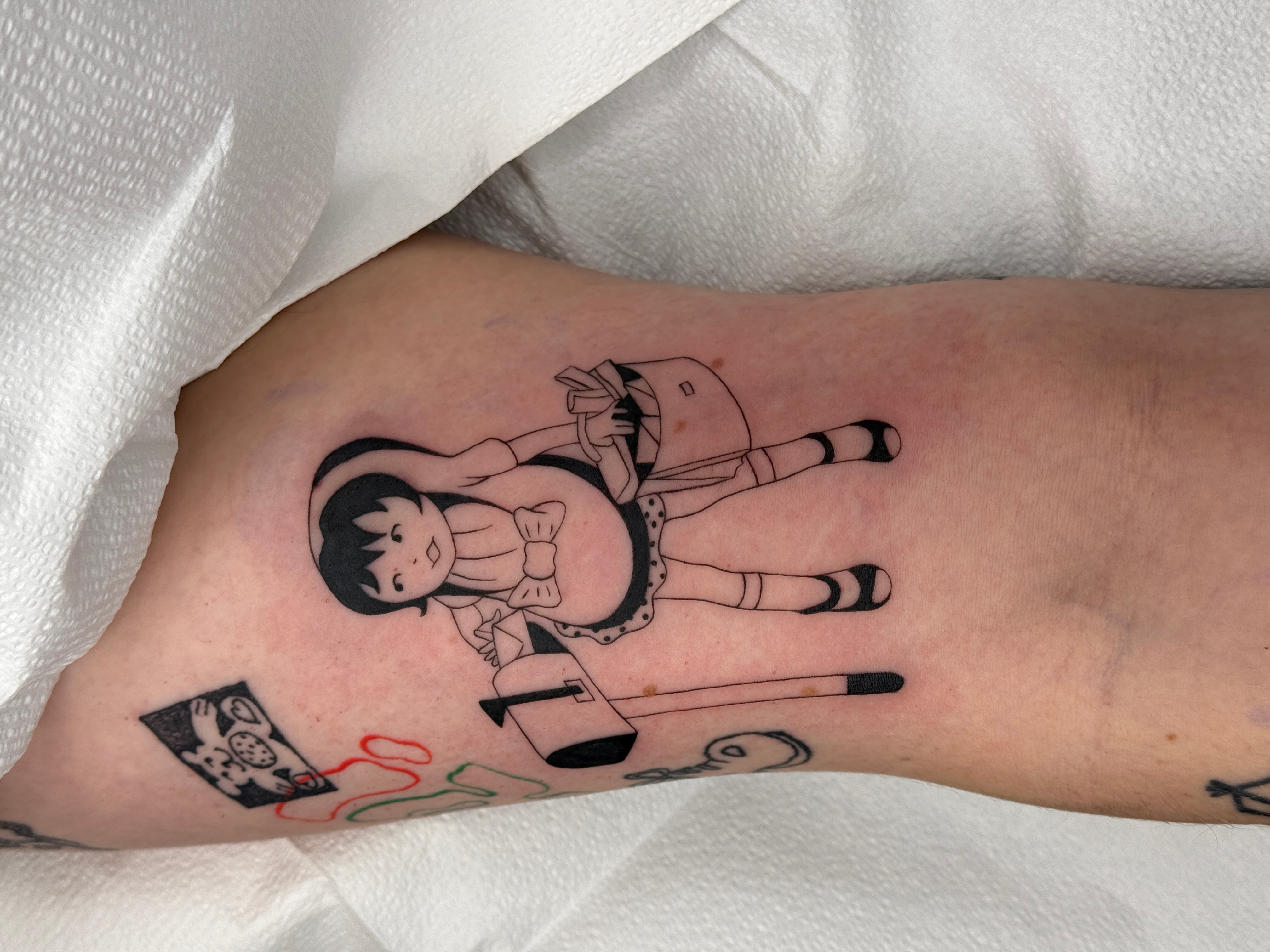Tattoo of a girl with short hair, wearing a dress, sitting with legs crossed, holding a stick, with a small flag nearby.