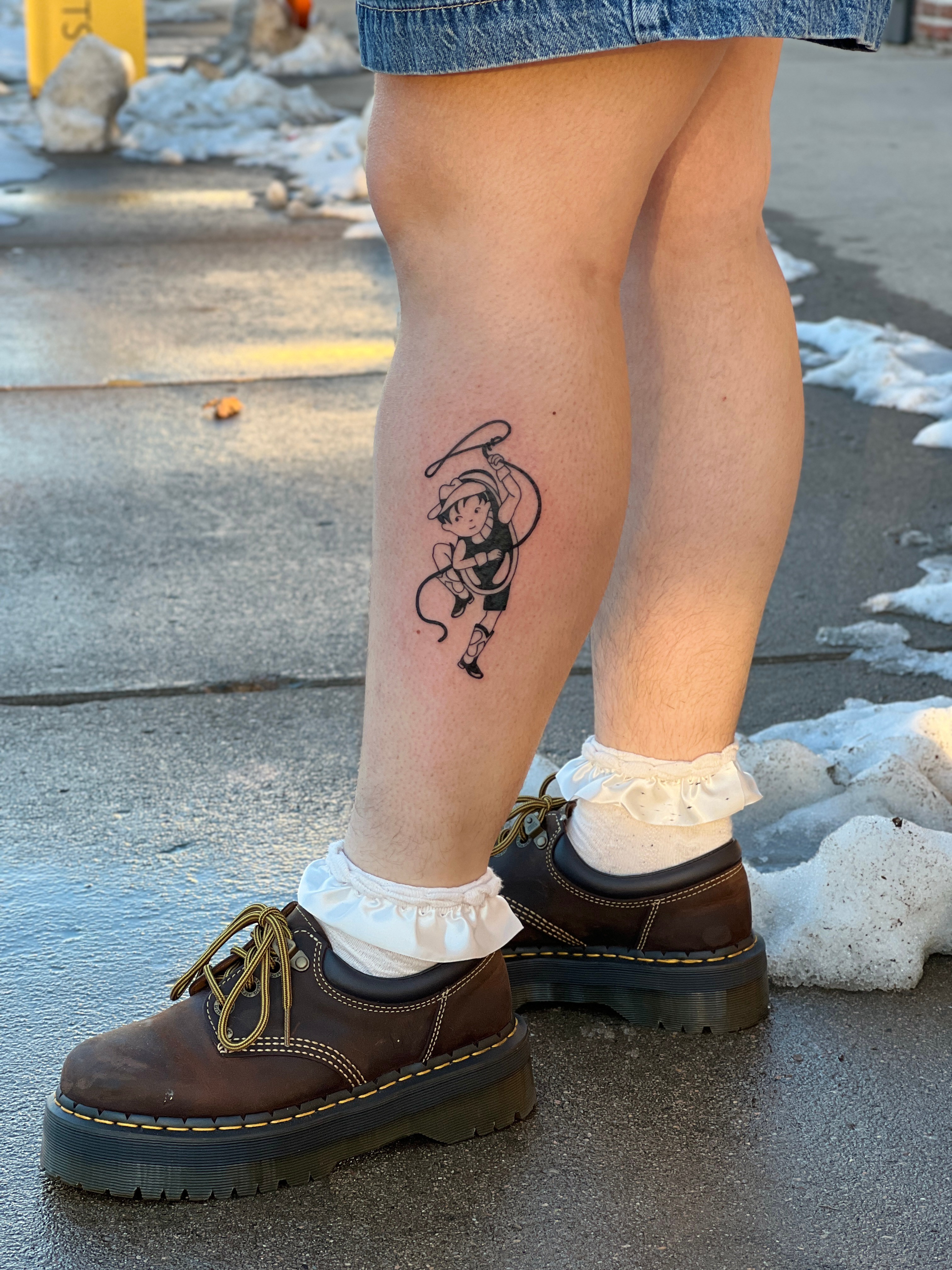 Person's lower legs and feet outdoors on wet pavement with snow, wearing brown shoes, white socks, and denim shorts, showing a tattoo of a cartoon character on the calf.