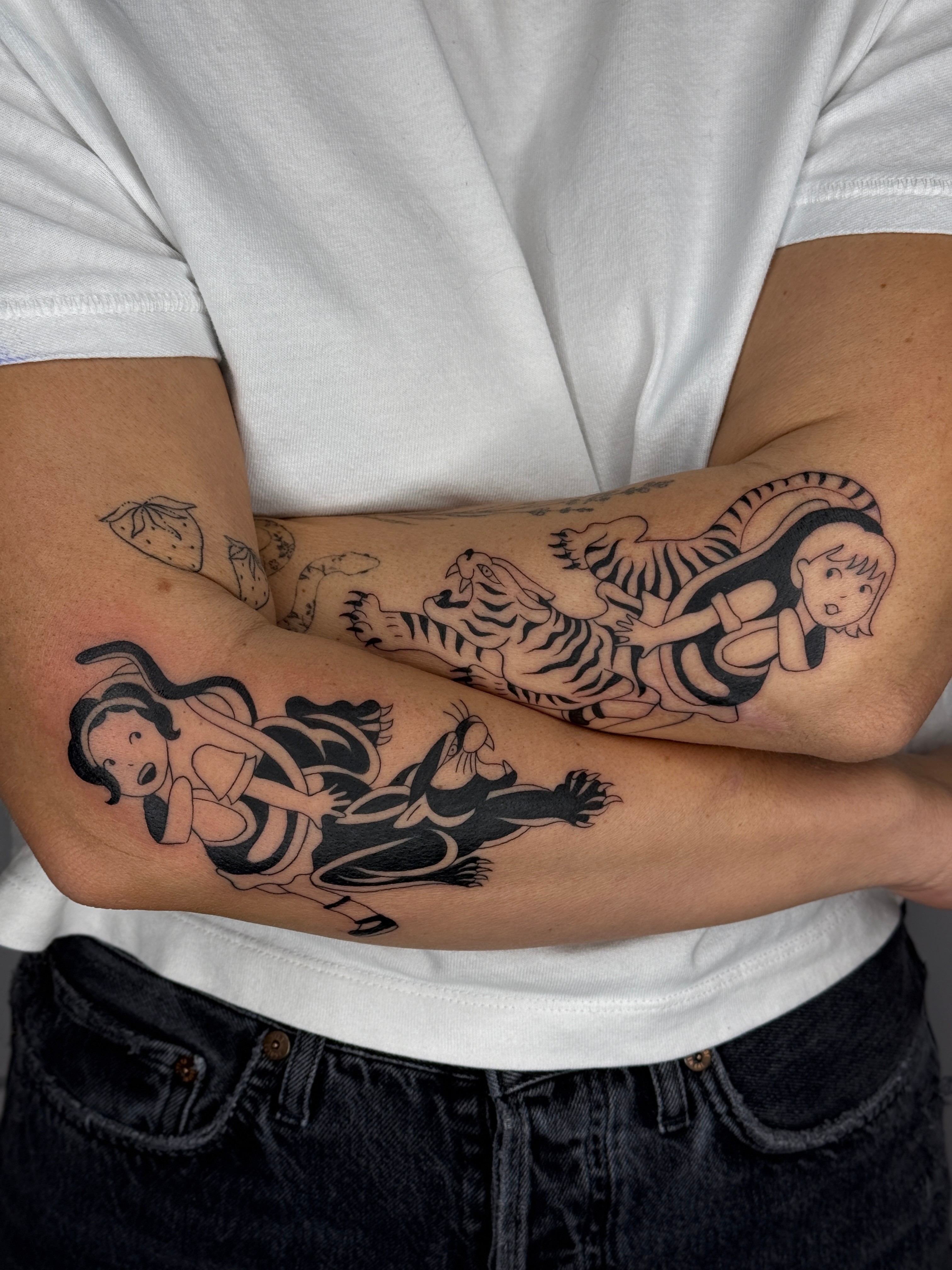Person with crossed arms showing tattoos of a mermaid and a tiger on forearms, wearing a white shirt and black pants.
