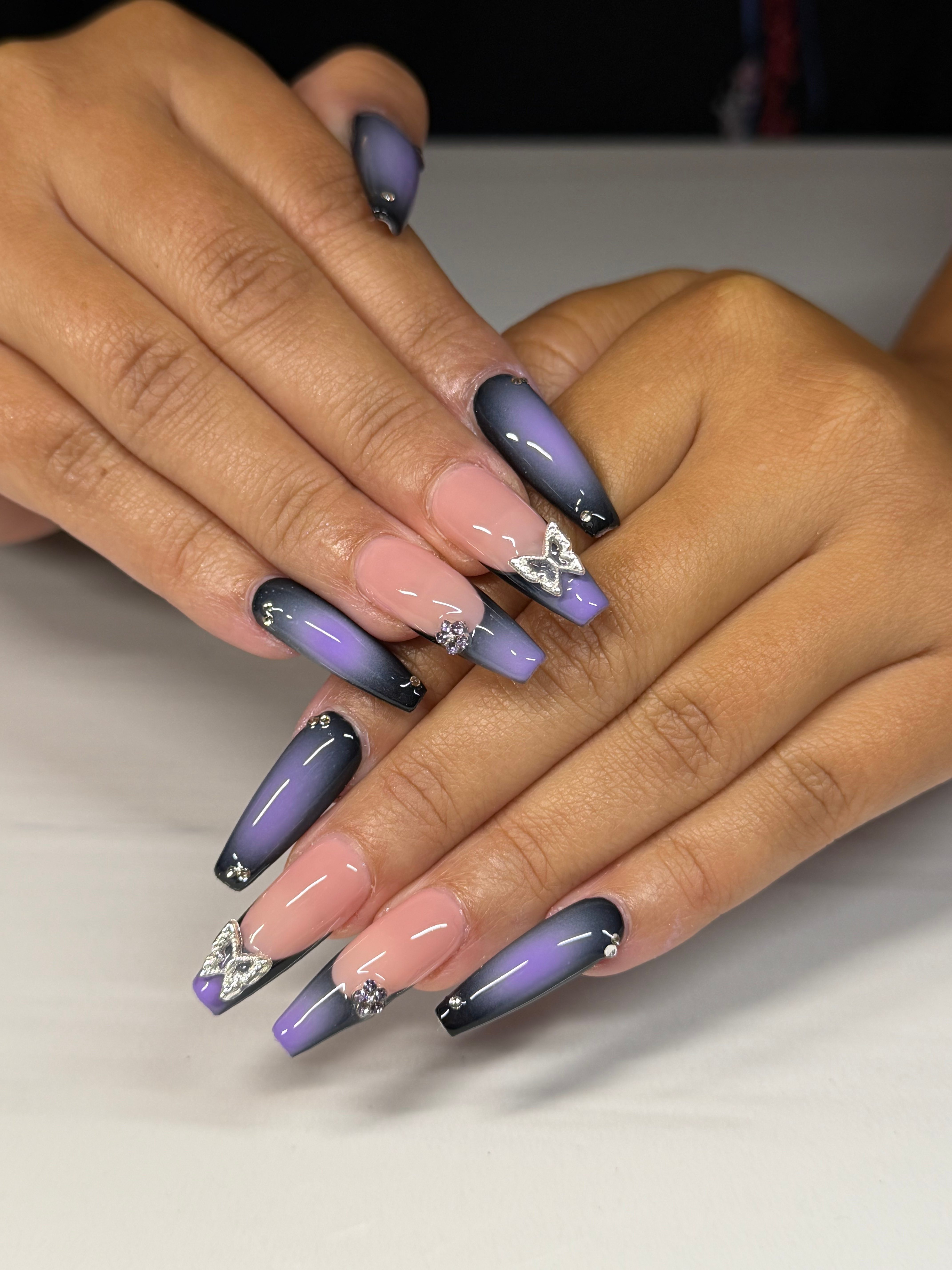 Close-up of hands with long, oval-shaped nails painted in gradient purple and black, with decorative rhinestones and designs.