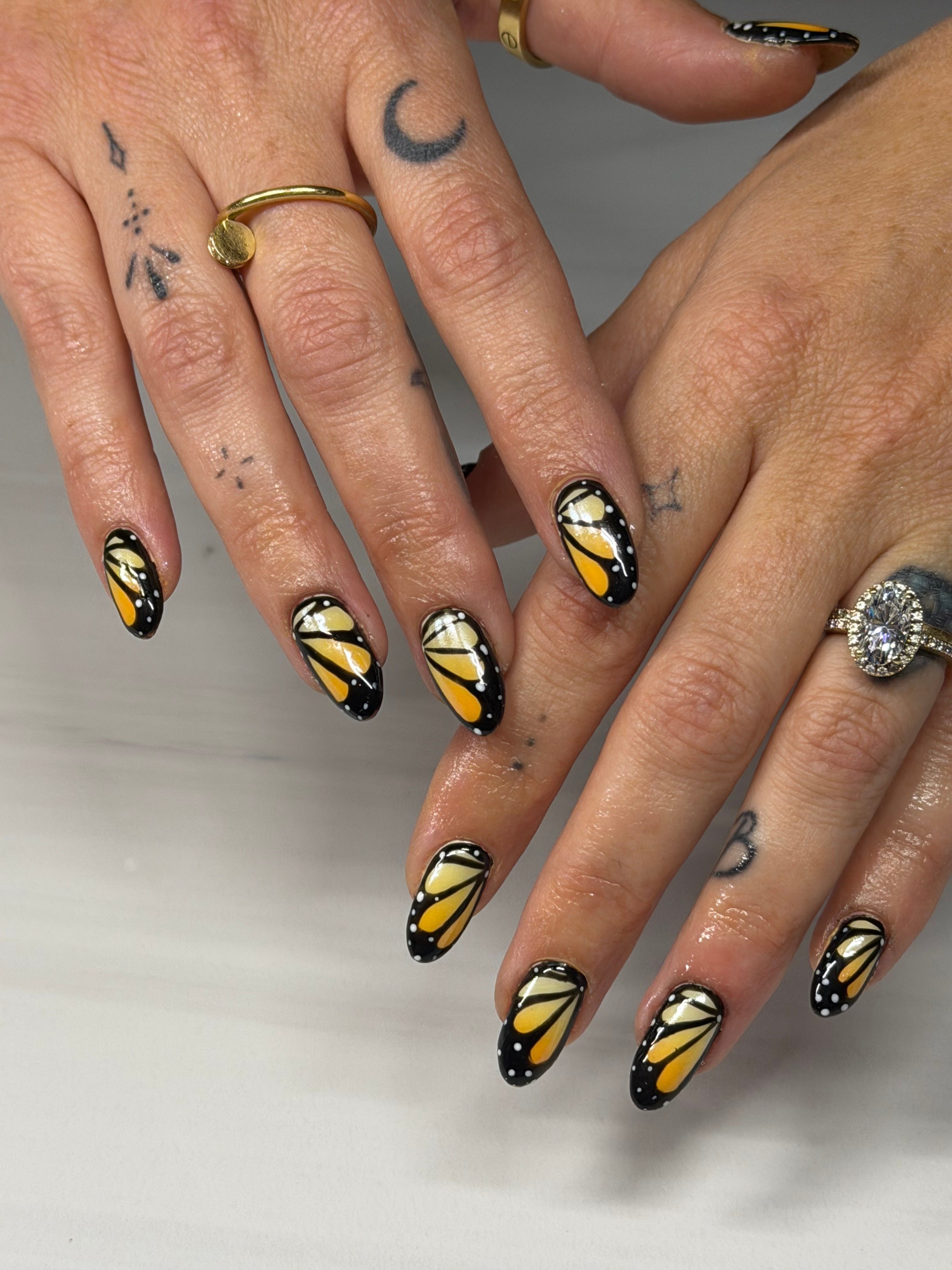 Two hands with butterfly nail art, rings, and tattoos, resting against a white surface.