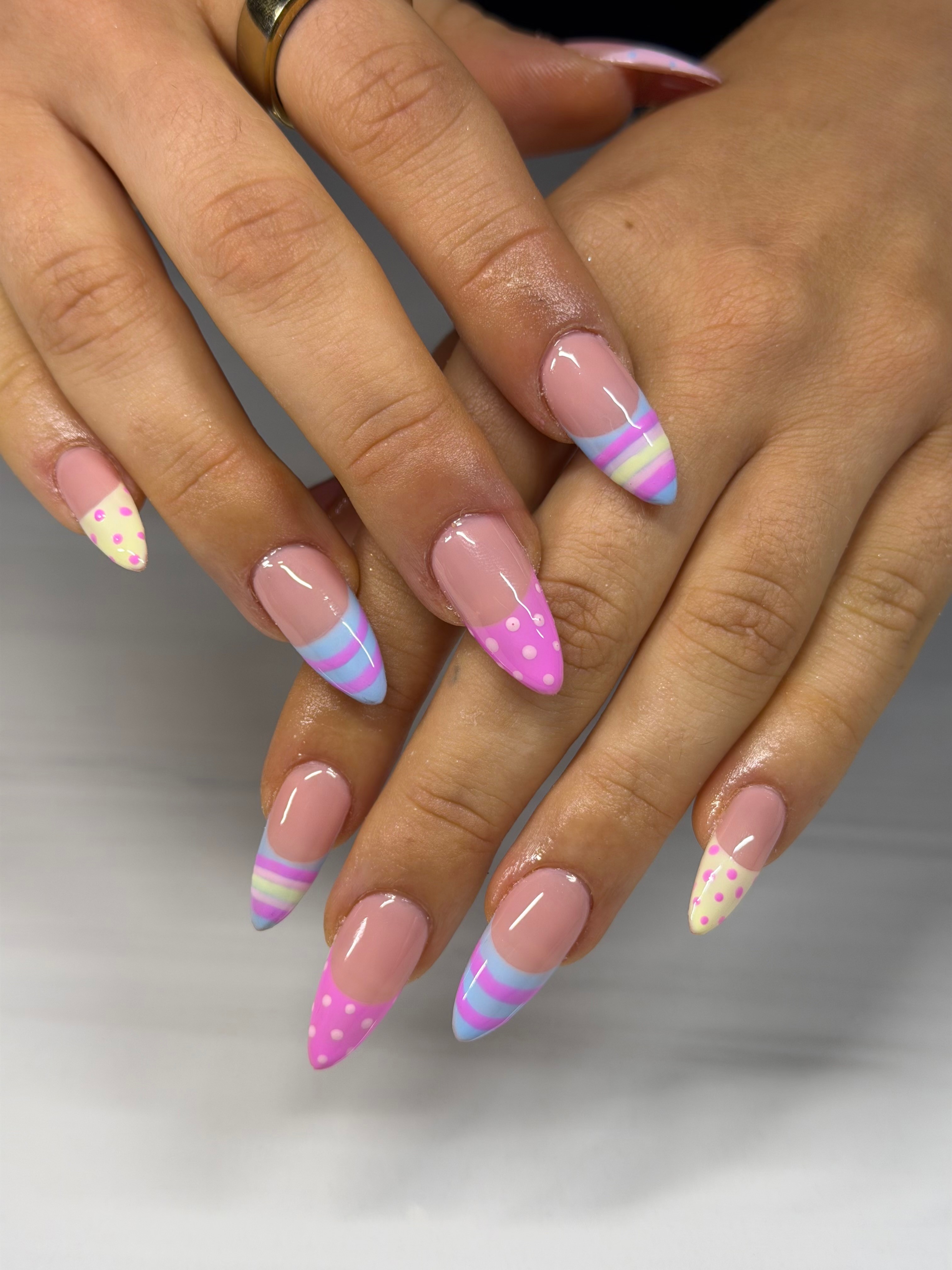 Hands with decorated nails featuring pink, purple, and white striped and polka dot designs, some with glitter accents.
