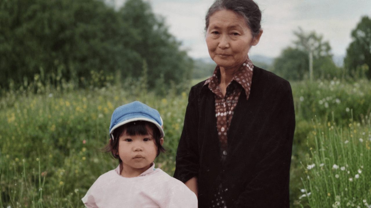 An elderly woman and a young girl stand outdoors in a field with greenery and flowers.