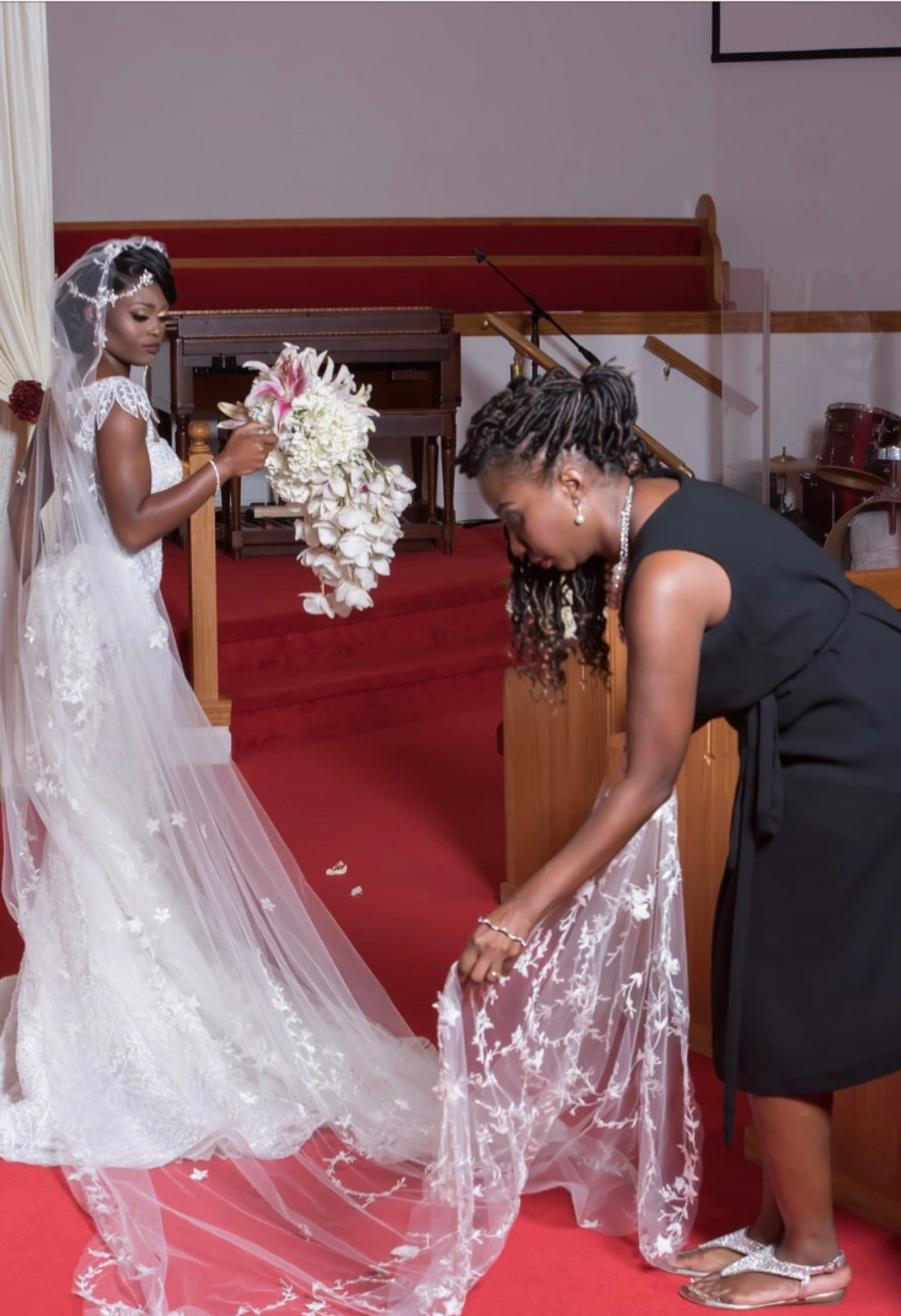 A bride in a white wedding dress holds a bouquet, while a woman adjusts her dress in a church interior.