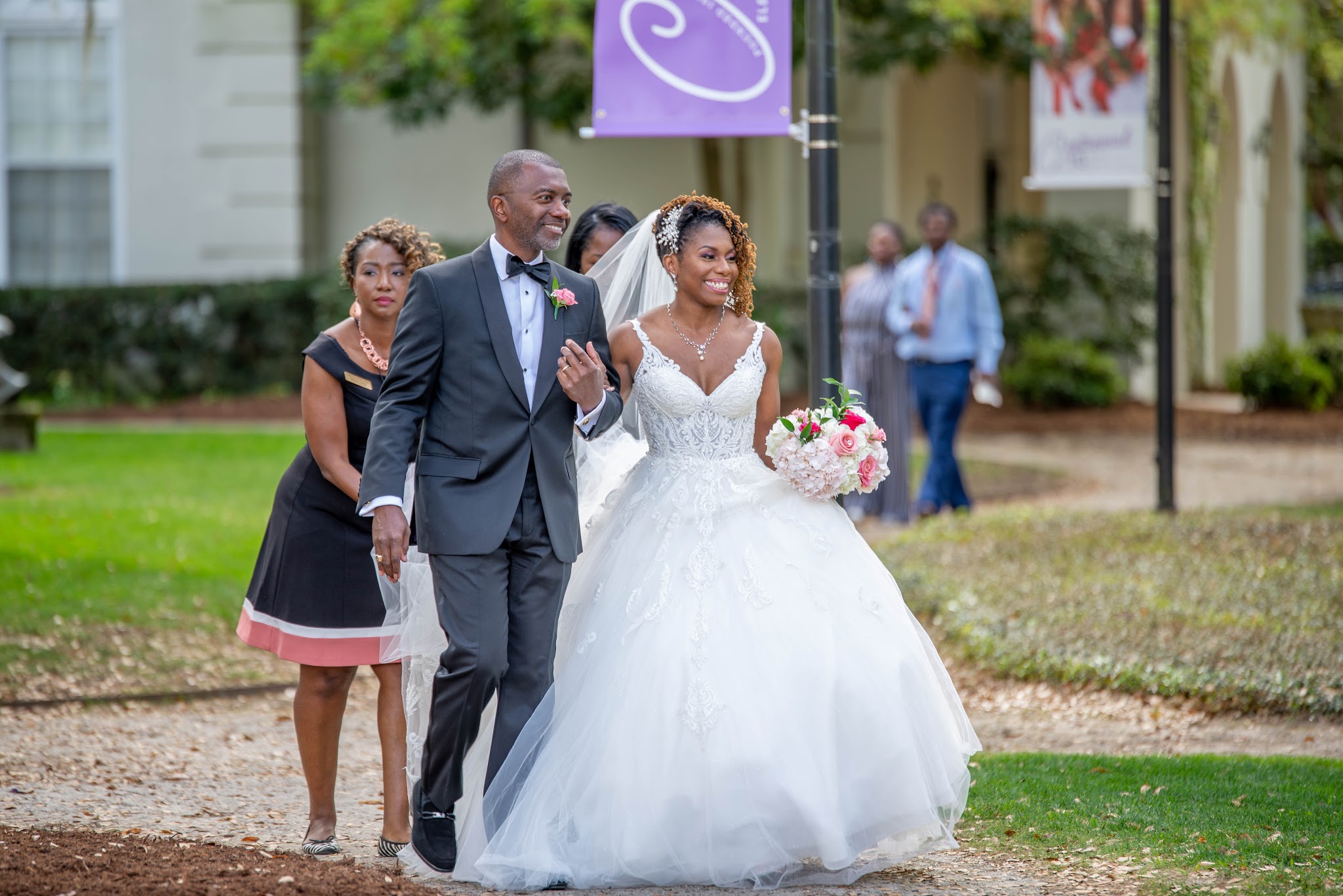 Bride in a white wedding dress holding a bouquet, walking with a man in a suit, outdoors on grass and pavement.