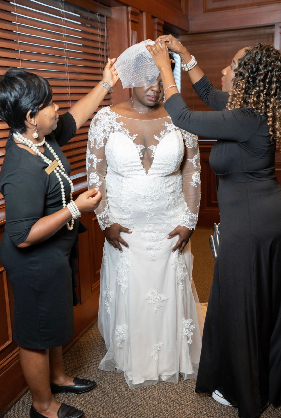 Three women in a room, one in a wedding dress, adjusting a veil on her head, wooden walls in background.