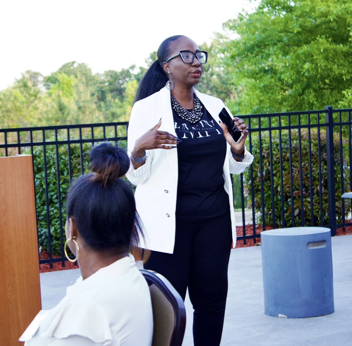 Woman speaking outdoors, holding a phone, with two seated women in foreground, green trees in background.