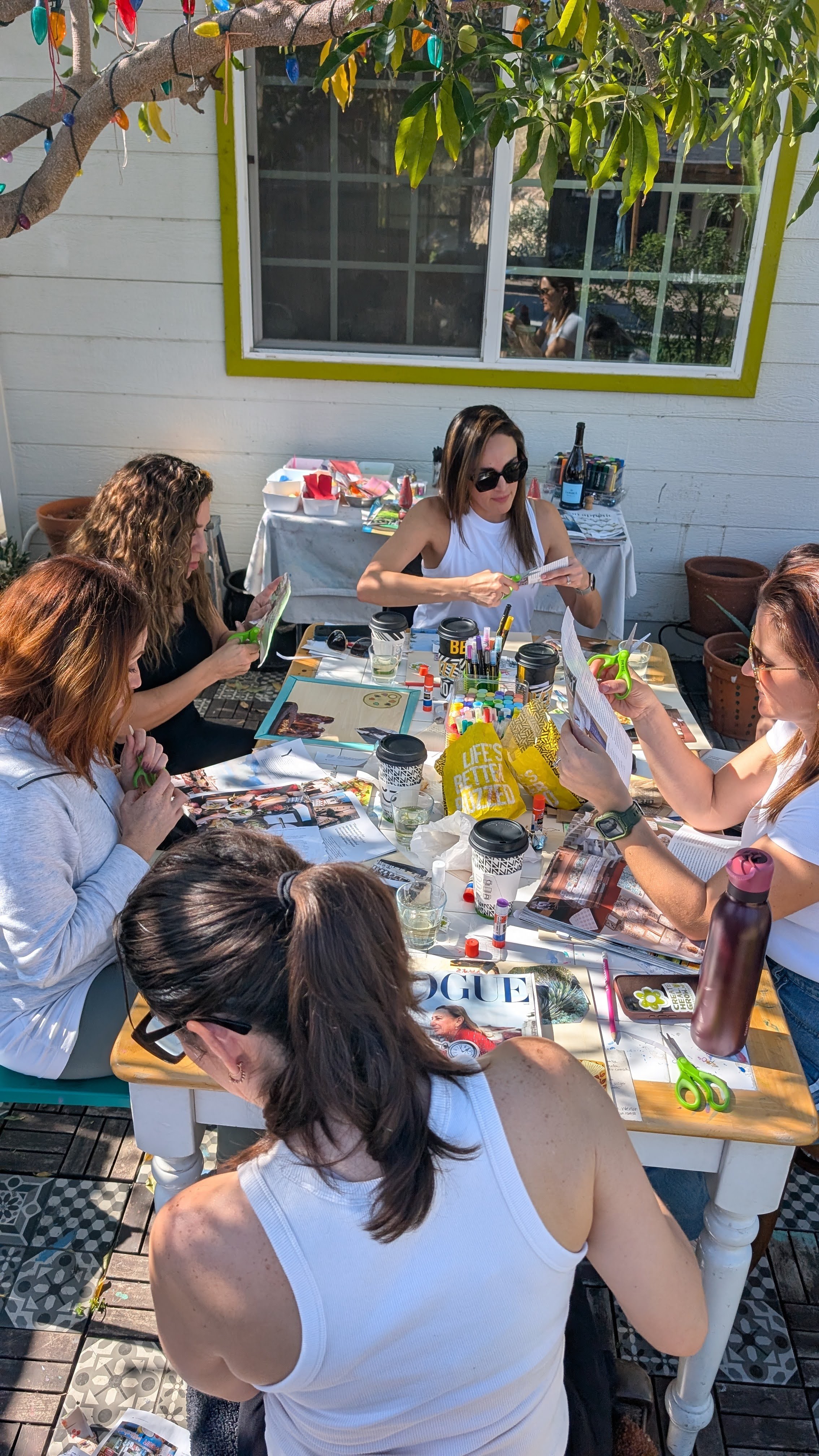 Group of women sitting around a table outdoors, enjoying drinks and conversation, with a white building and window in background.