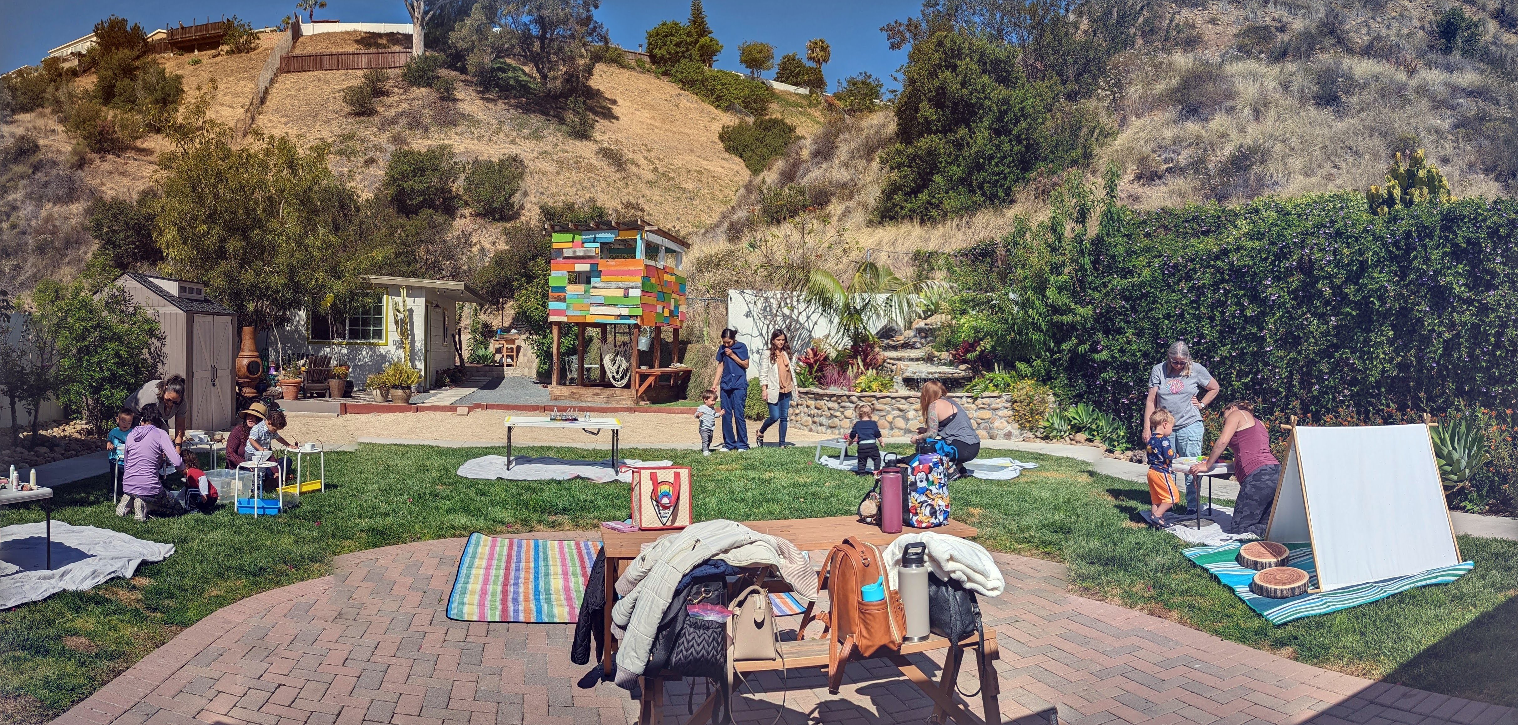 People gather outdoors in a park with trees, hills, and buildings in the background, some sitting and others standing.