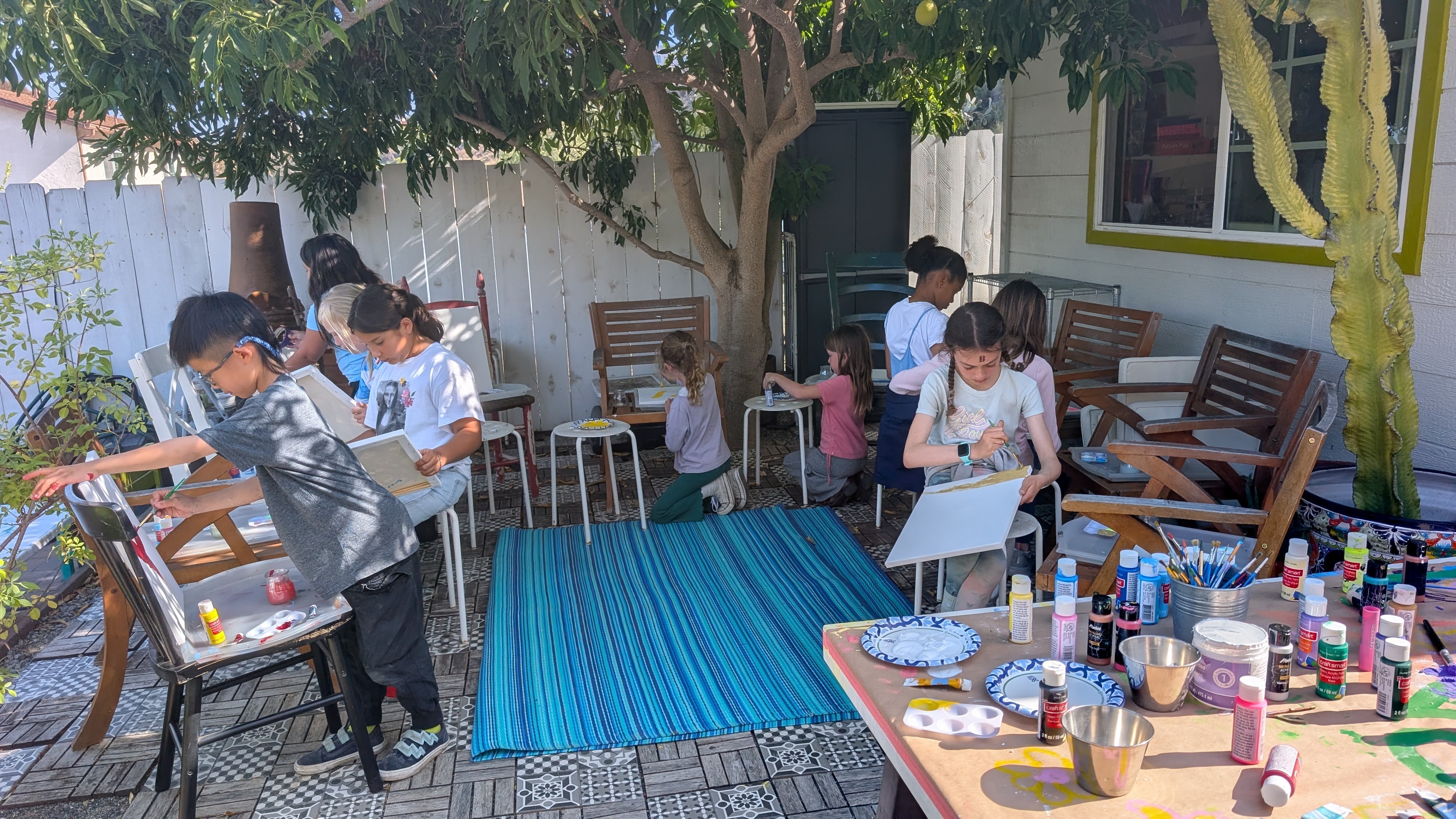 Children painting outdoors on a patio with trees and a white fence, with art supplies on a table nearby.