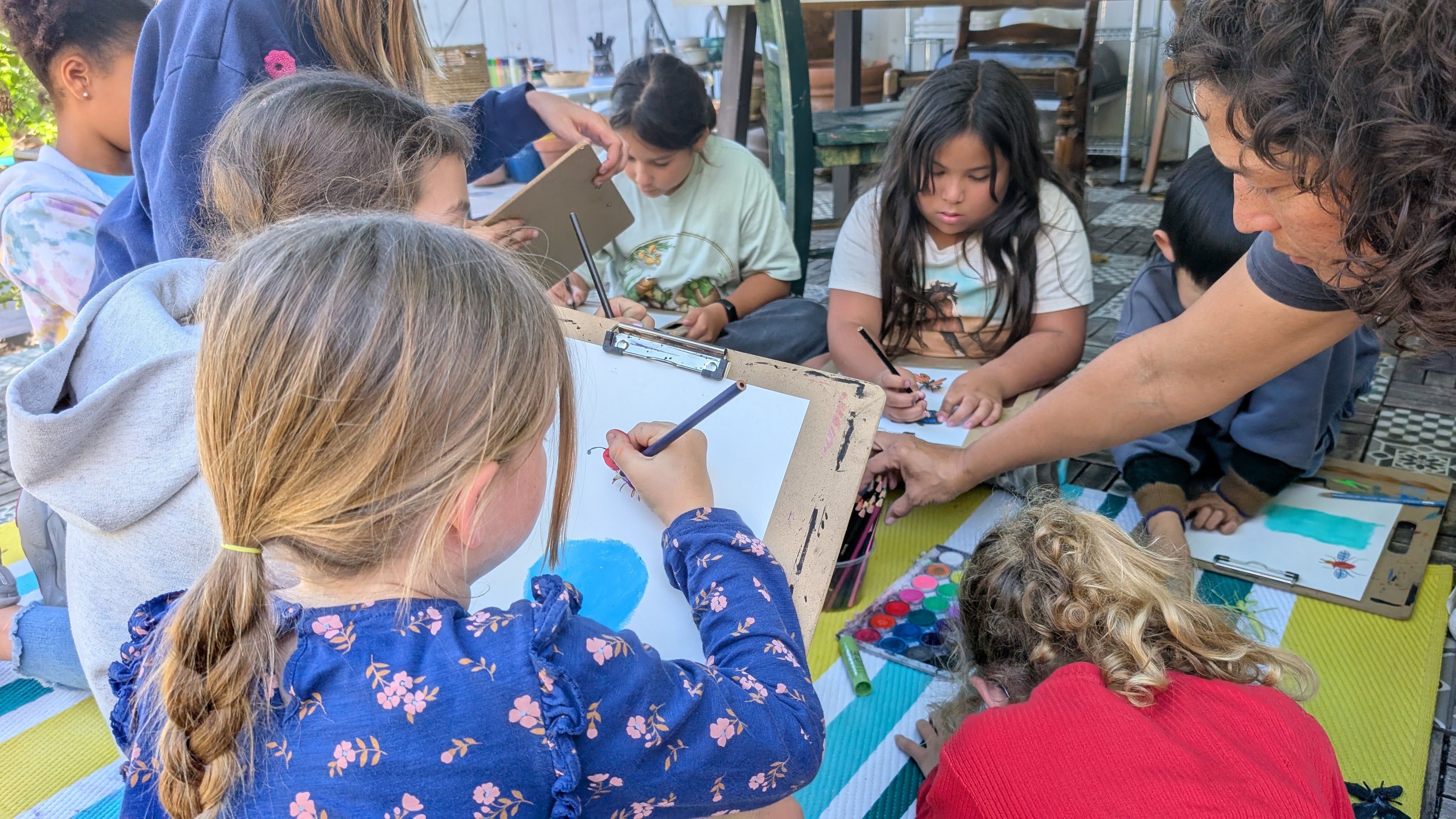 Children sit around a table drawing and coloring with markers and colored pencils, supervised by an adult. Bright classroom setting.