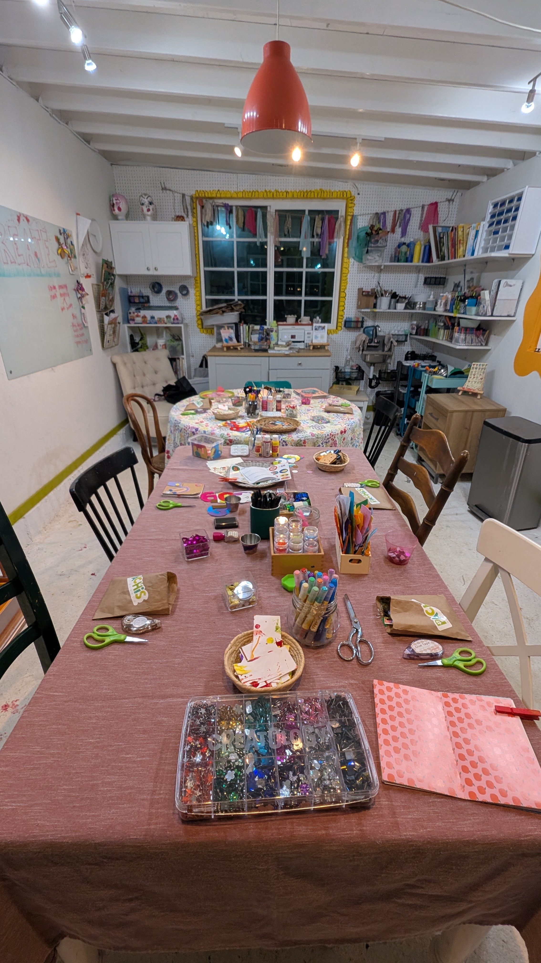 Party table with pink tablecloth, decorated with gifts, plates, and scissors, in a room with shelves and a window.