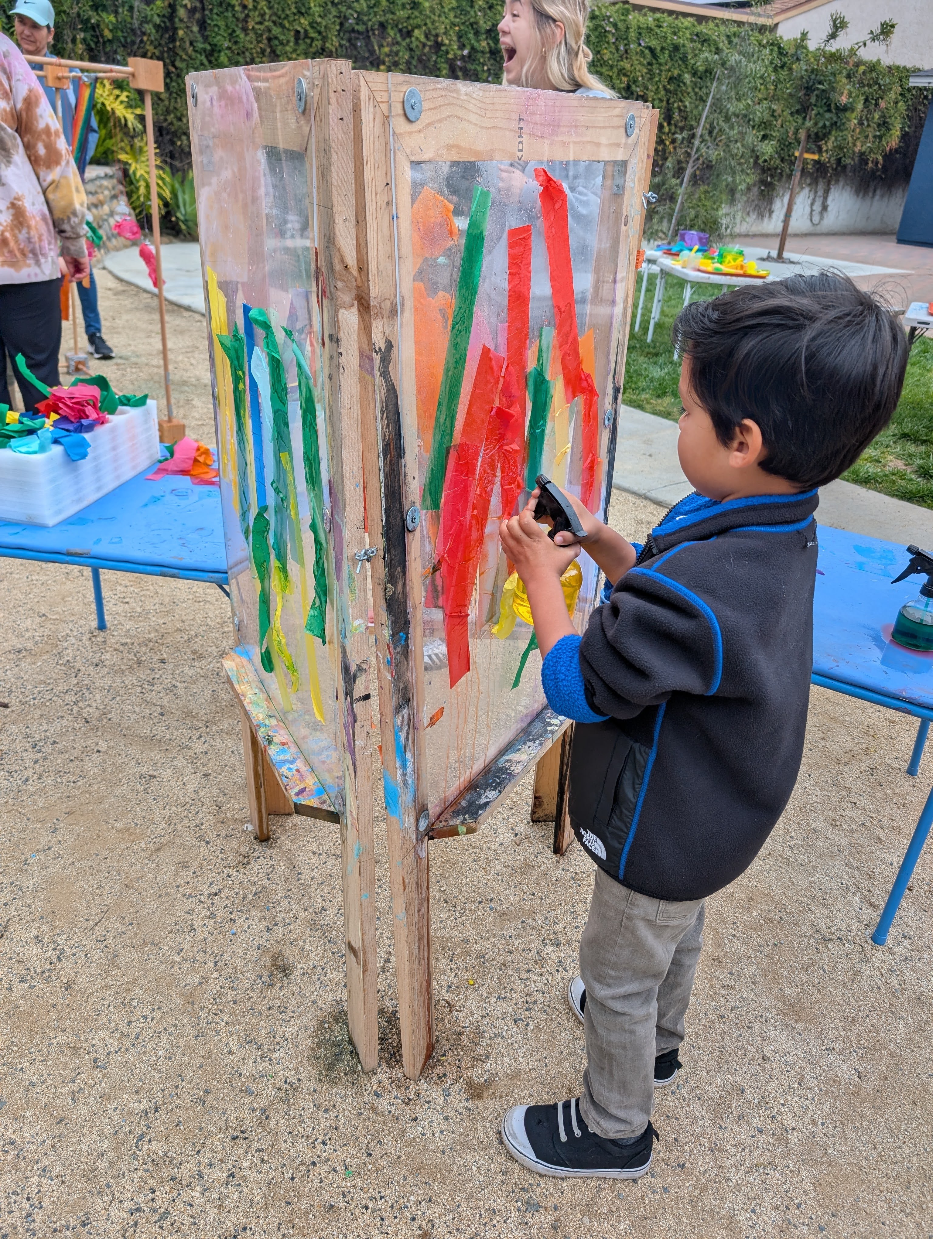 Child painting on a wooden easel outdoors, with other children and tables in the background.