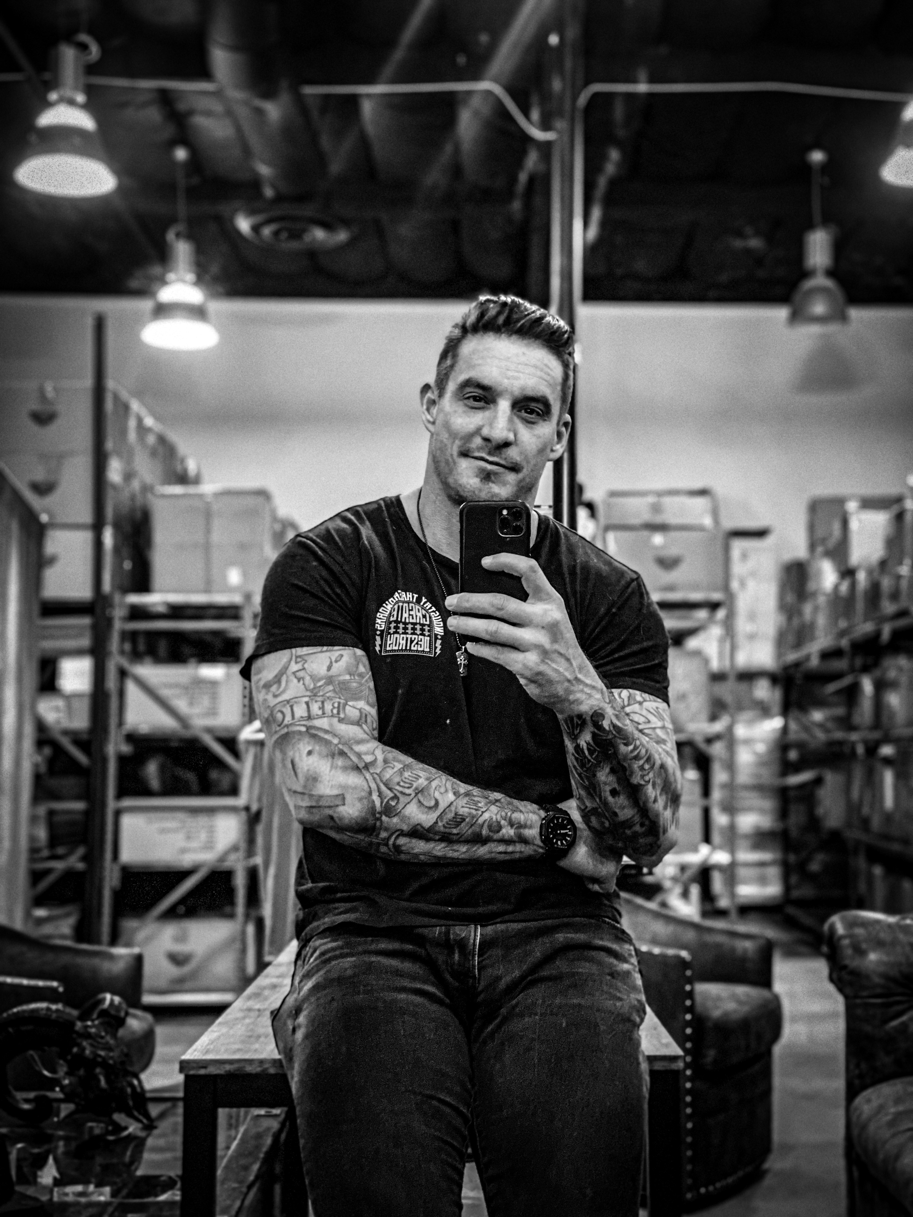 Man with tattoos taking a selfie in a warehouse or industrial space, smiling, with shelves and equipment in background.