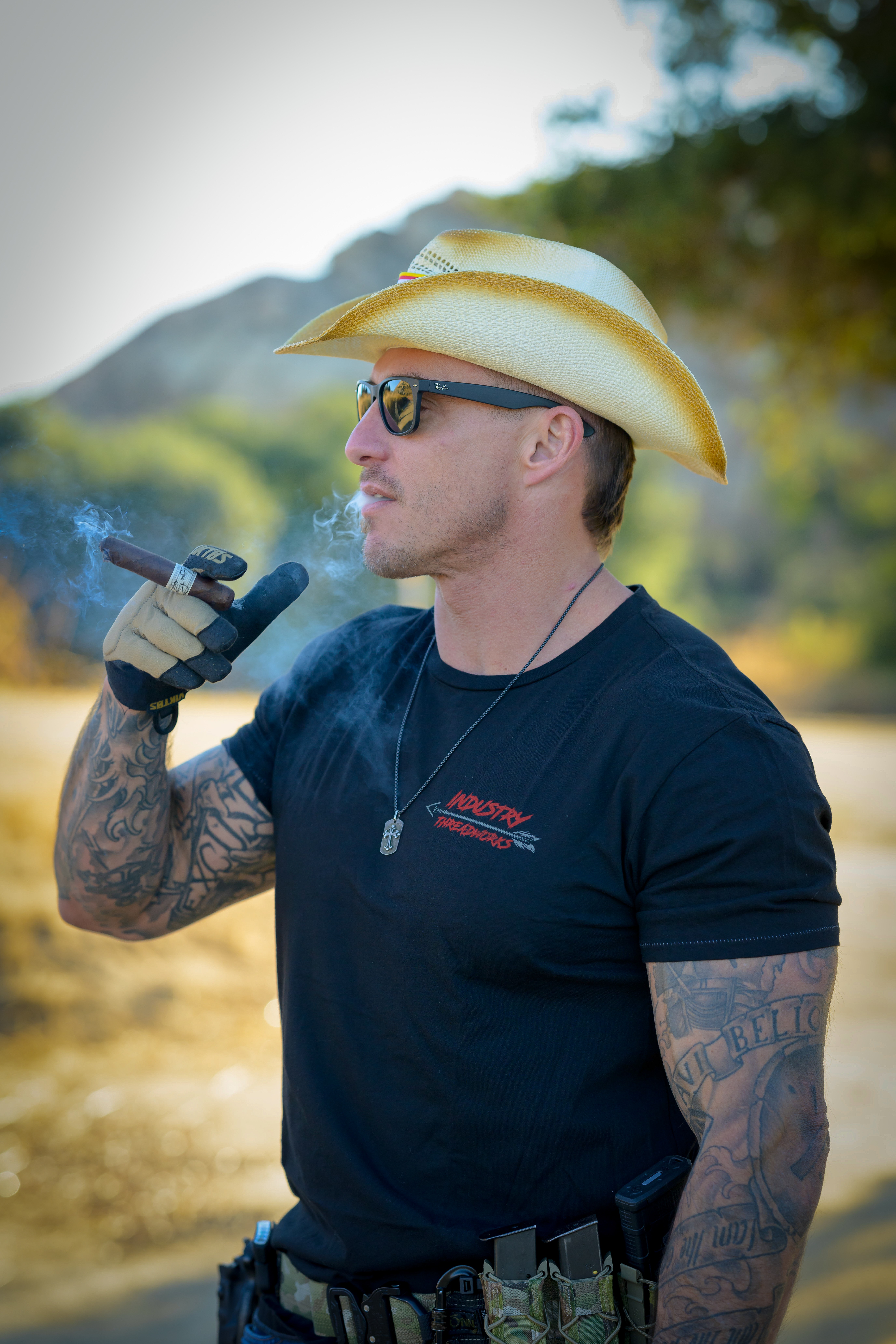 Man wearing a cowboy hat and sunglasses, smoking a pipe outdoors with mountains and trees in background.