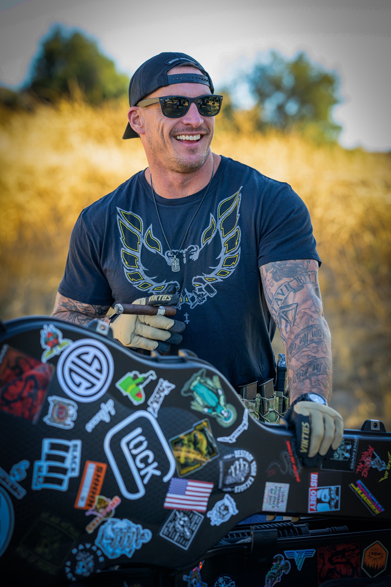 Smiling man wearing sunglasses, a cap, and a graphic t-shirt outdoors, with a tattooed arm and a decorated motorcycle.