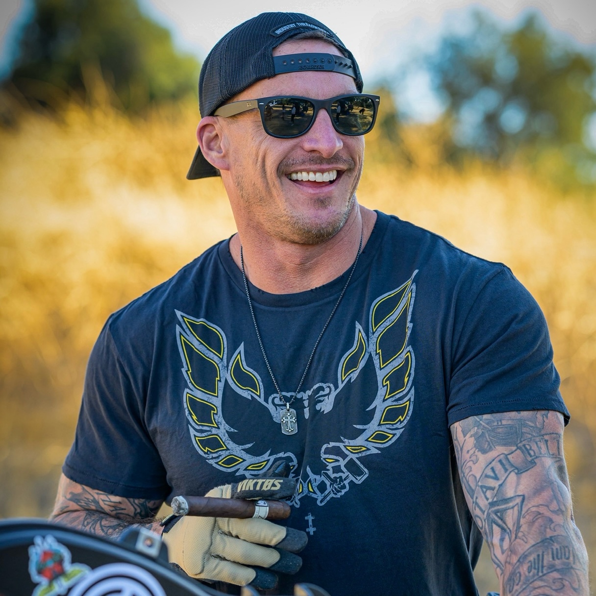 Smiling man wearing sunglasses, a black cap backward, a dark T-shirt with a butterfly design, and tattoos, outdoors.