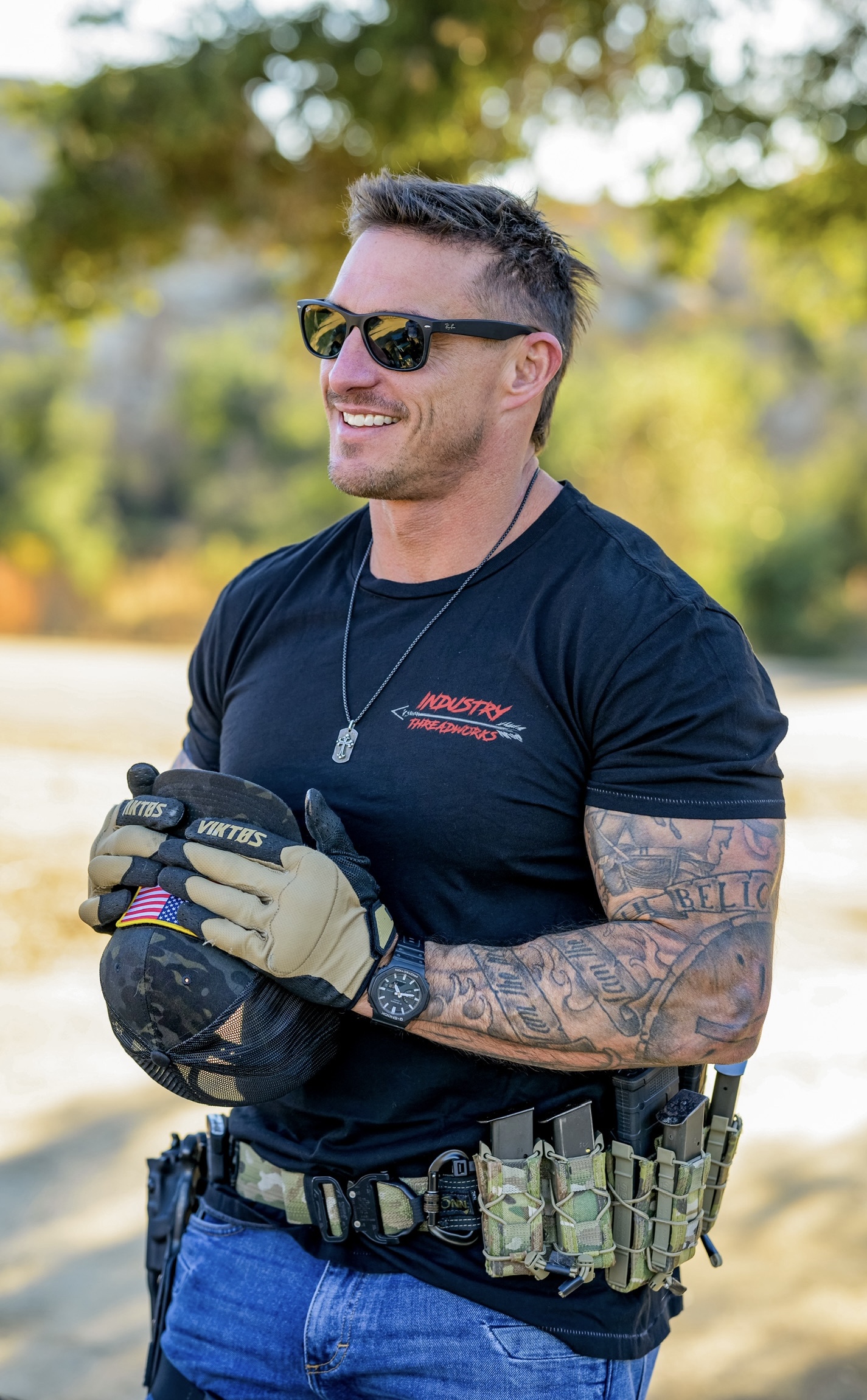 Smiling man wearing sunglasses, black shirt, gloves, and a utility belt outdoors with trees in background.