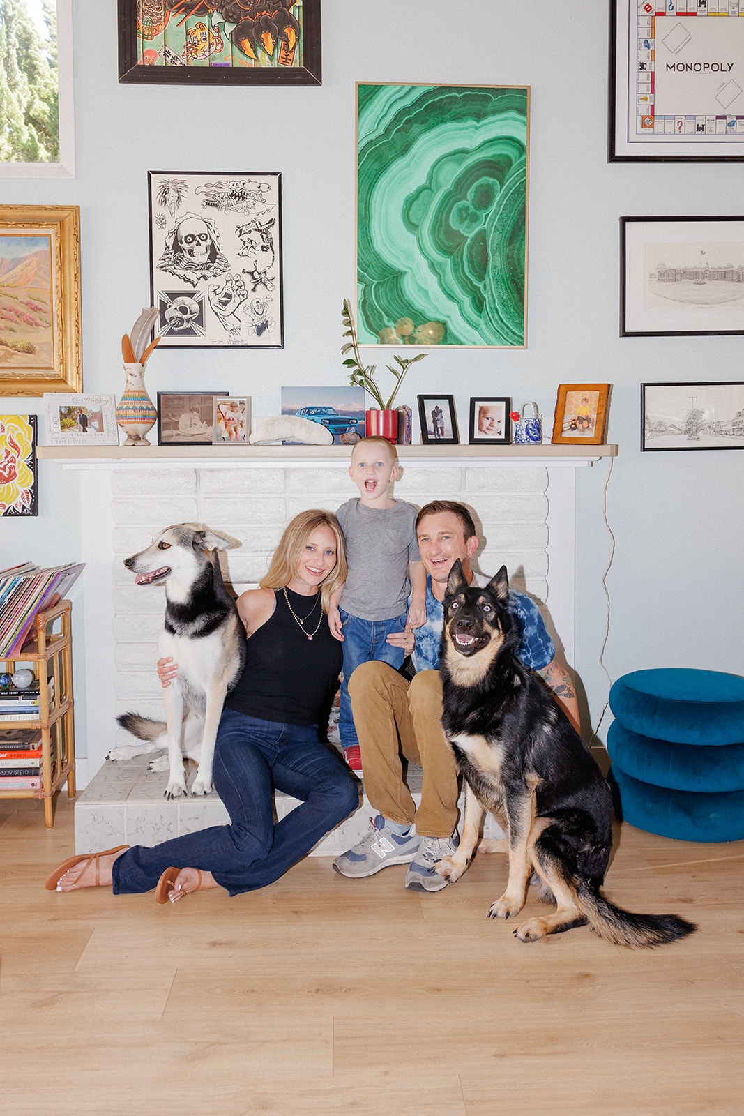 Three people with two dogs in a living room with colorful artwork on the wall, smiling and sitting on the floor.