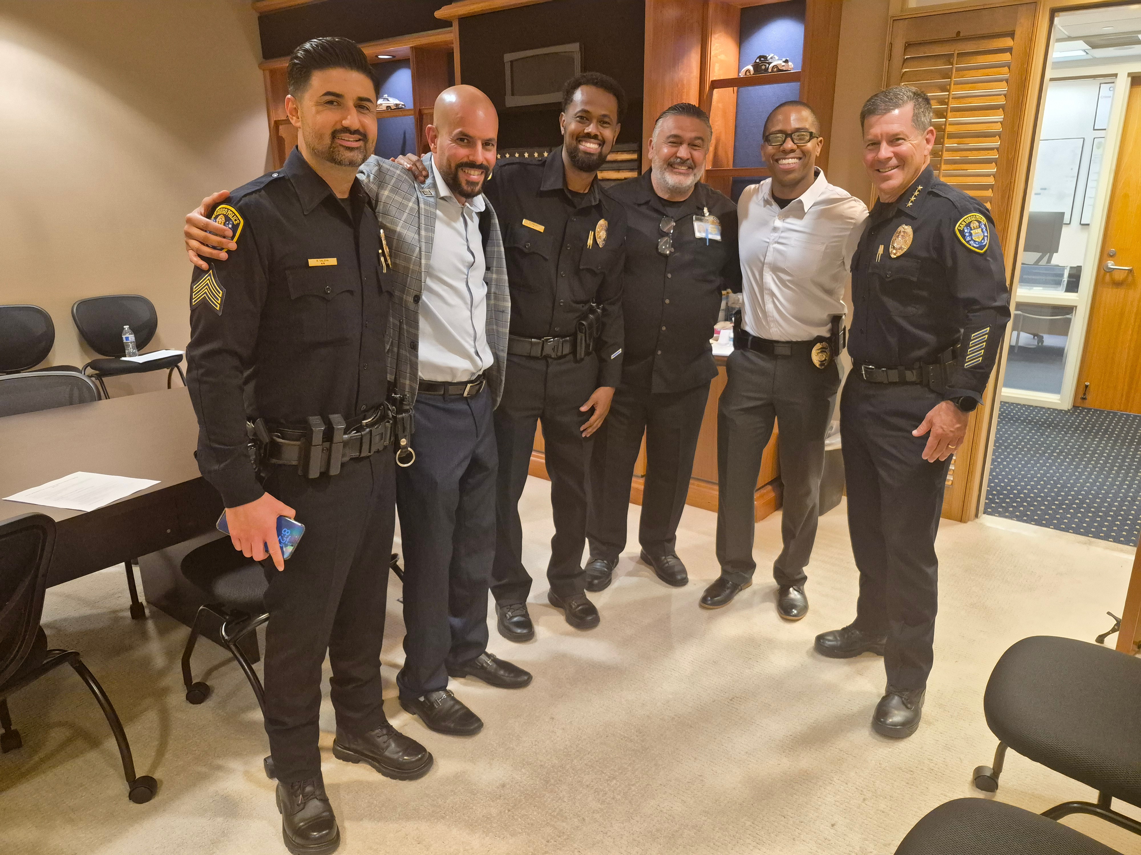 Six men, some in police uniforms and others in civilian clothing, stand together indoors smiling. They are in a room with chairs and wooden panels.