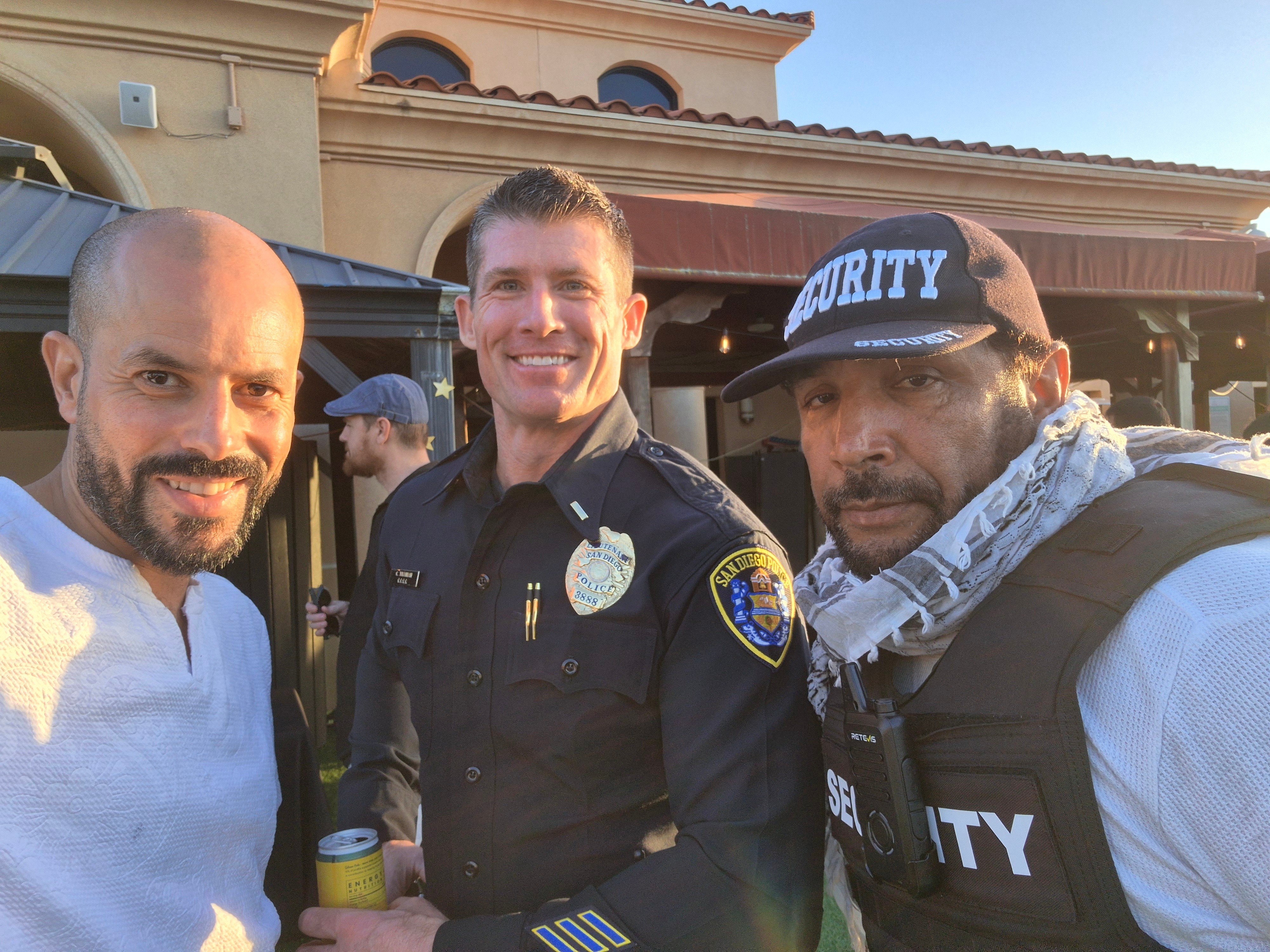 Three men, including a police officer, smiling outdoors in front of a building, with two men wearing security vests.