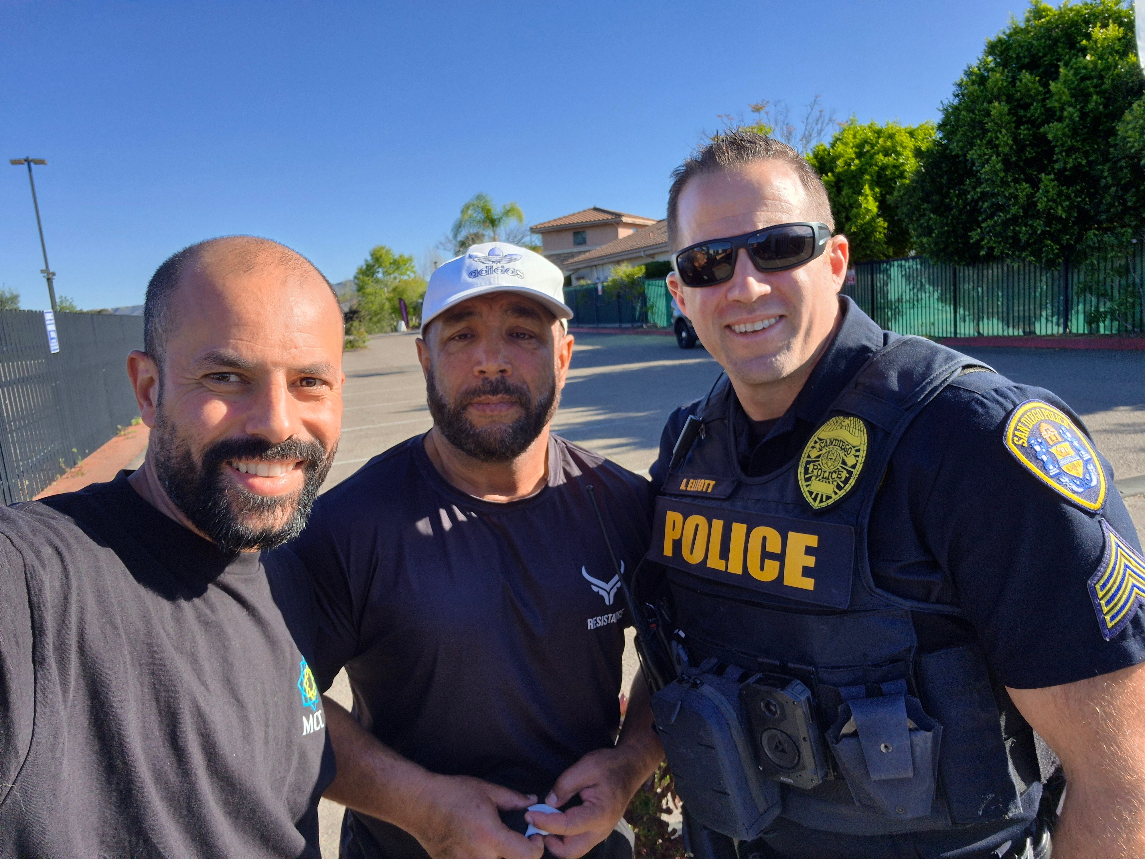 Three men standing outdoors, one in police uniform, smiling at the camera, with trees and a building in the background.