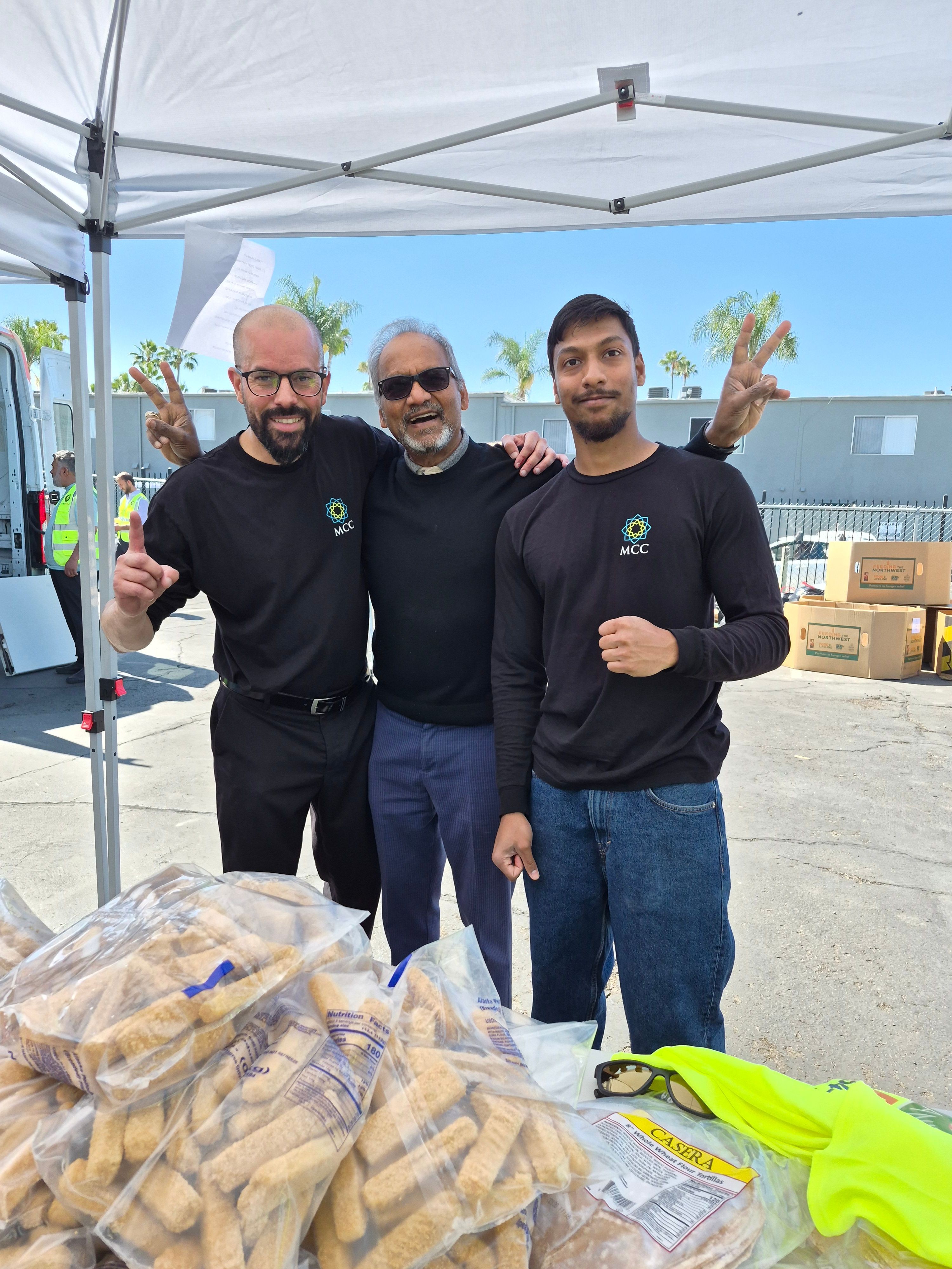 Three men standing outdoors under a tent, smiling and waving, with a table and yellow safety vest nearby.