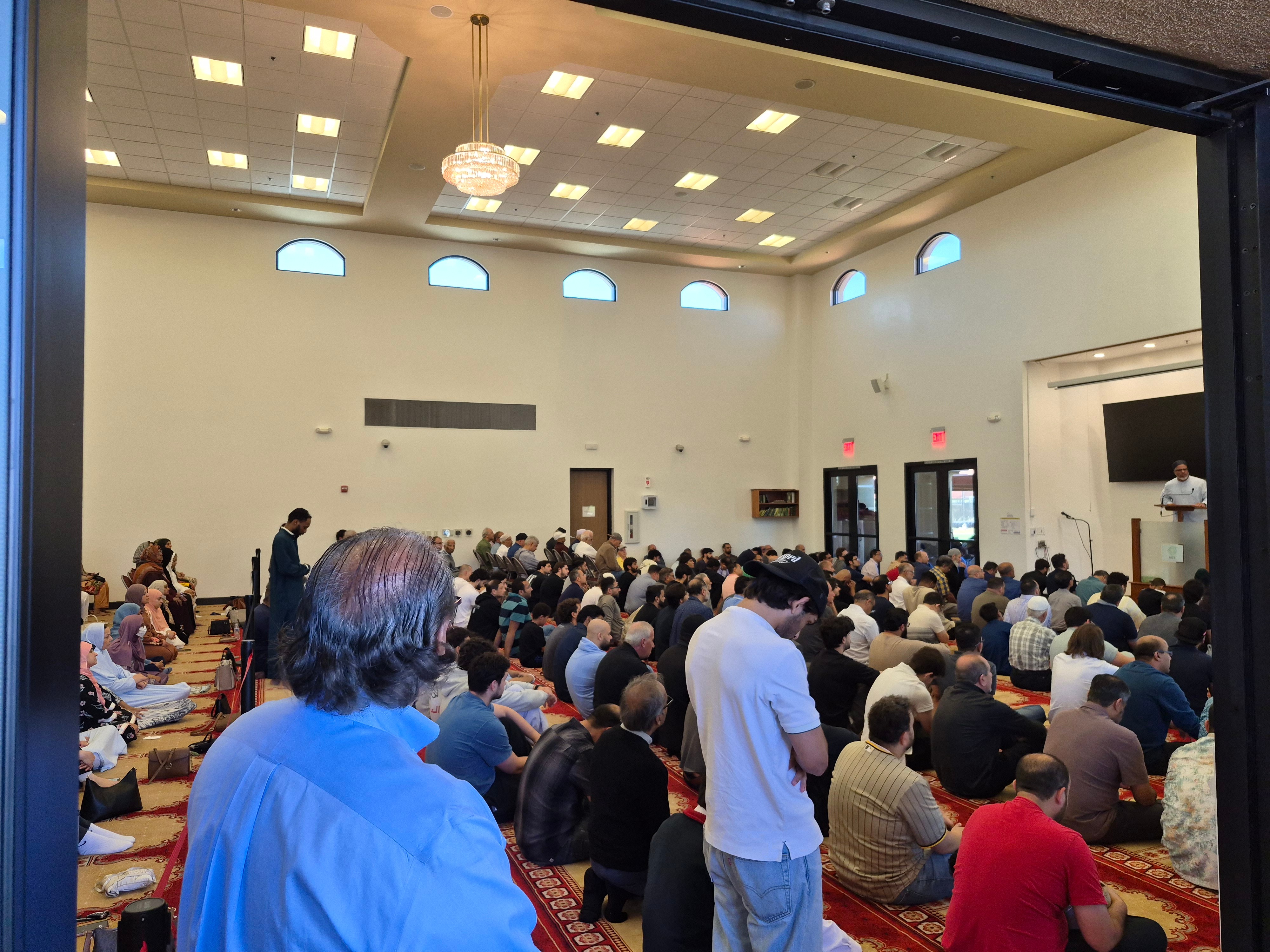 Large group of people seated in a spacious room with high ceiling, windows, and a chandelier, attending an event or presentation.