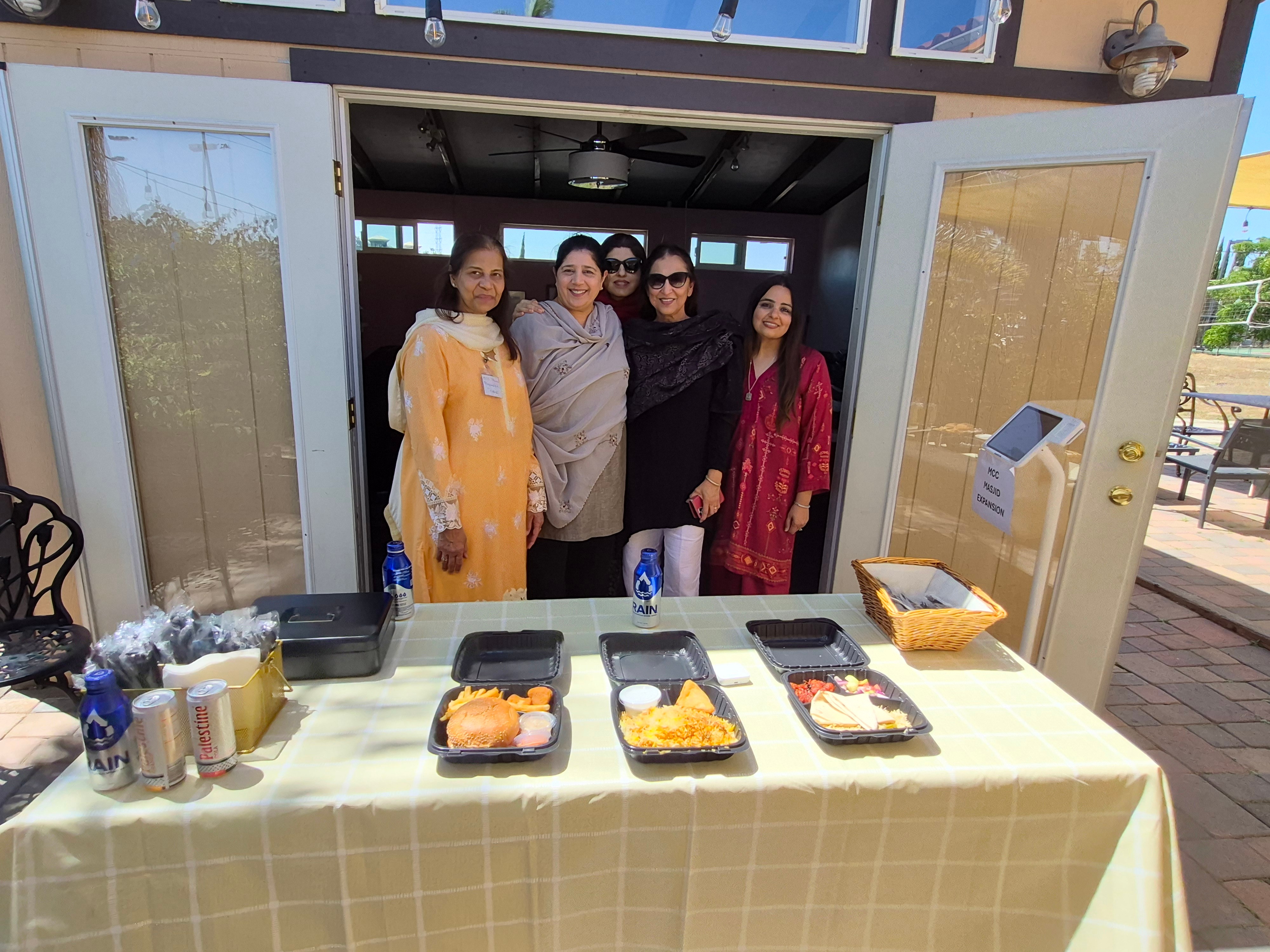 Four women stand behind a table with food trays outdoors, open doors behind them, sunny day, some chairs visible.