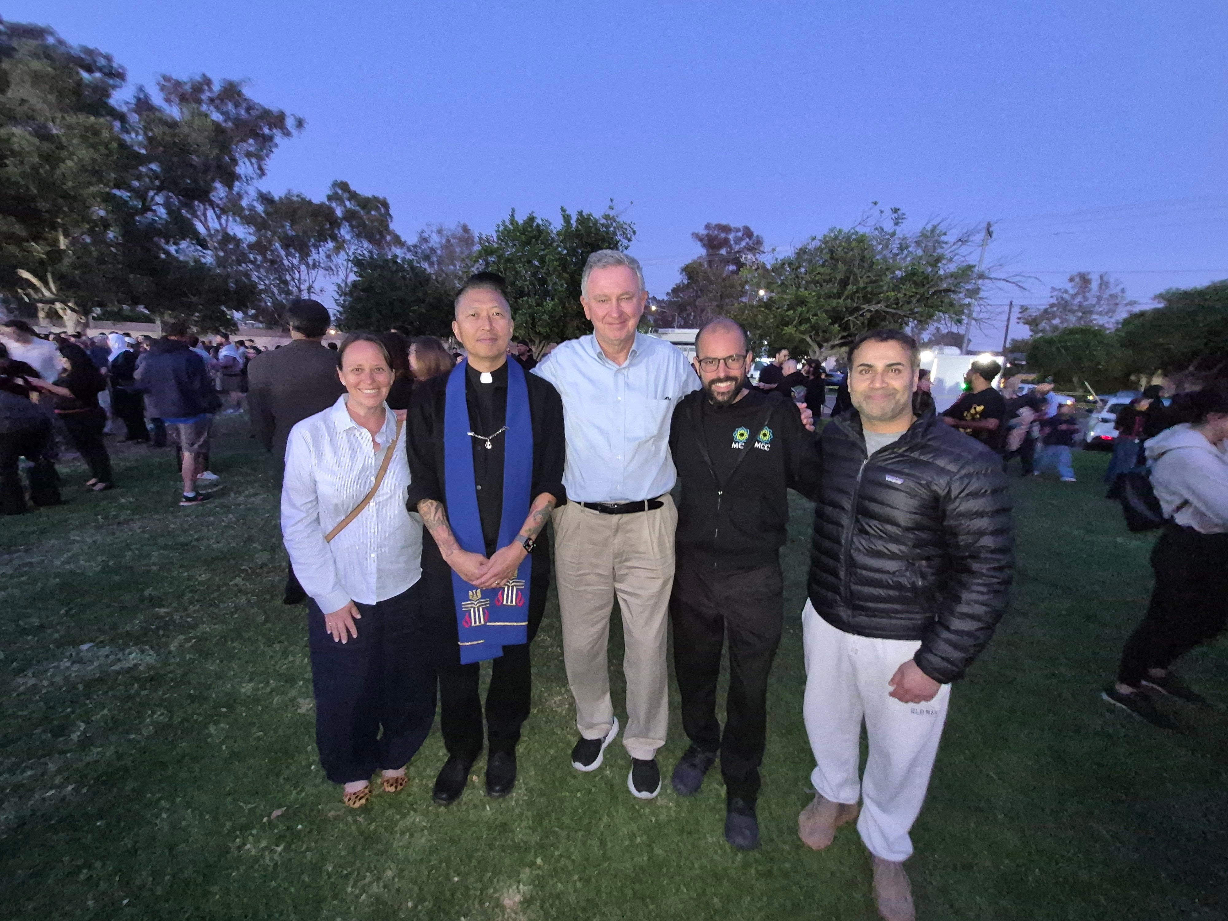 Five people standing outdoors in a grassy area with trees and a crowd in the background.
