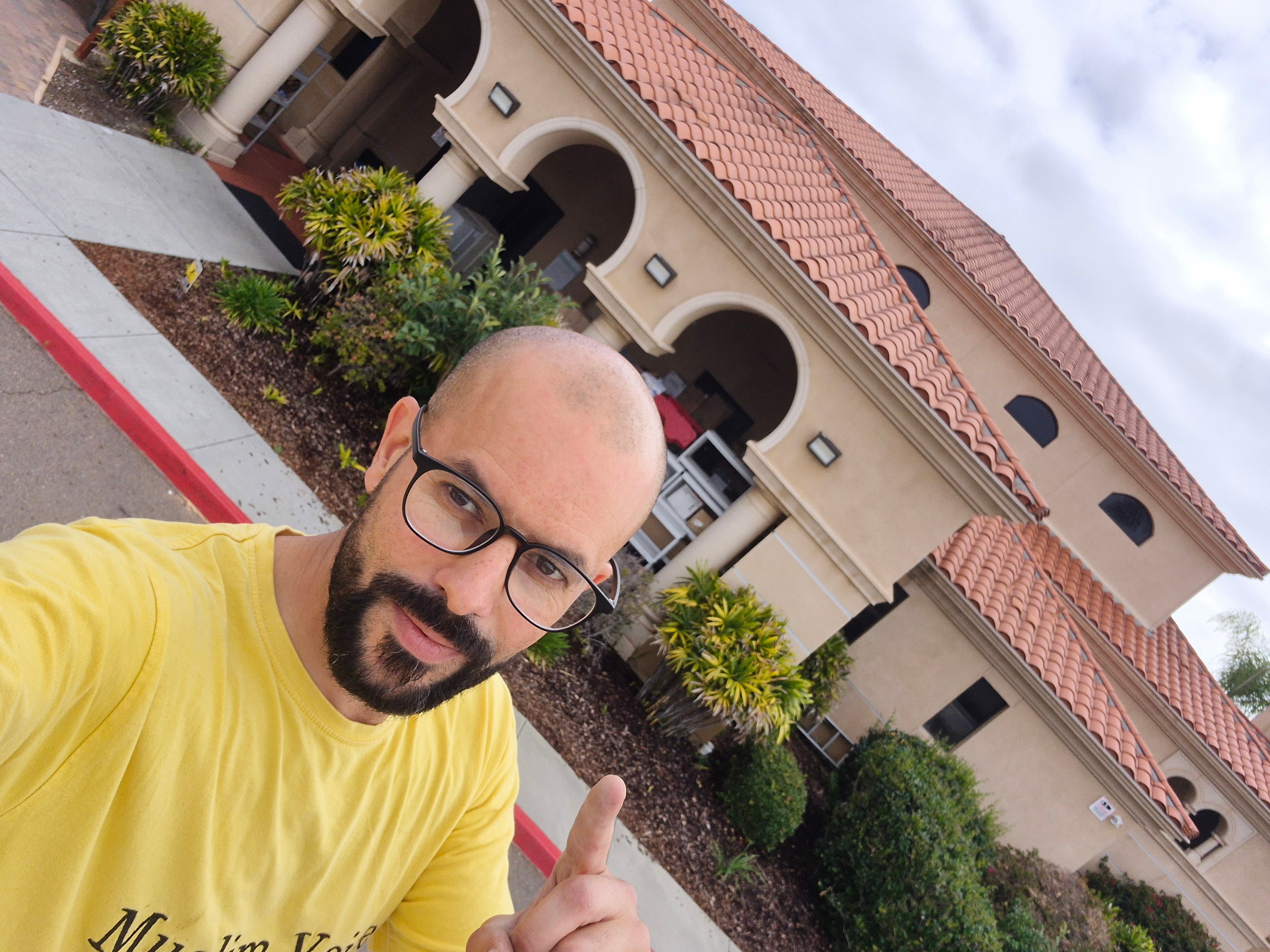 Man with glasses and a beard wearing a yellow shirt taking a selfie in front of a building with arches and a red-tiled roof.