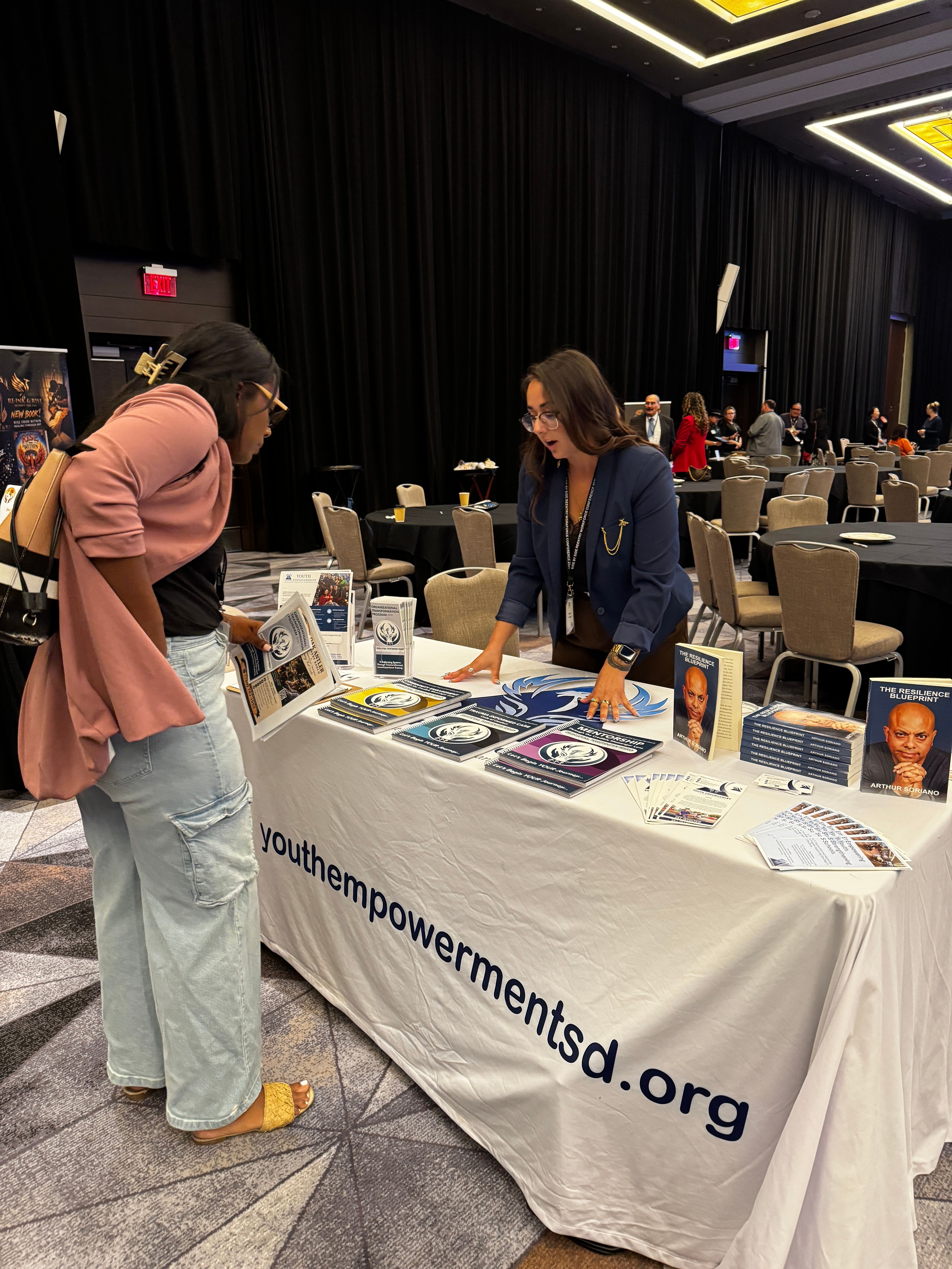 Two women talking at a table with informational materials, in a conference or exhibition setting.