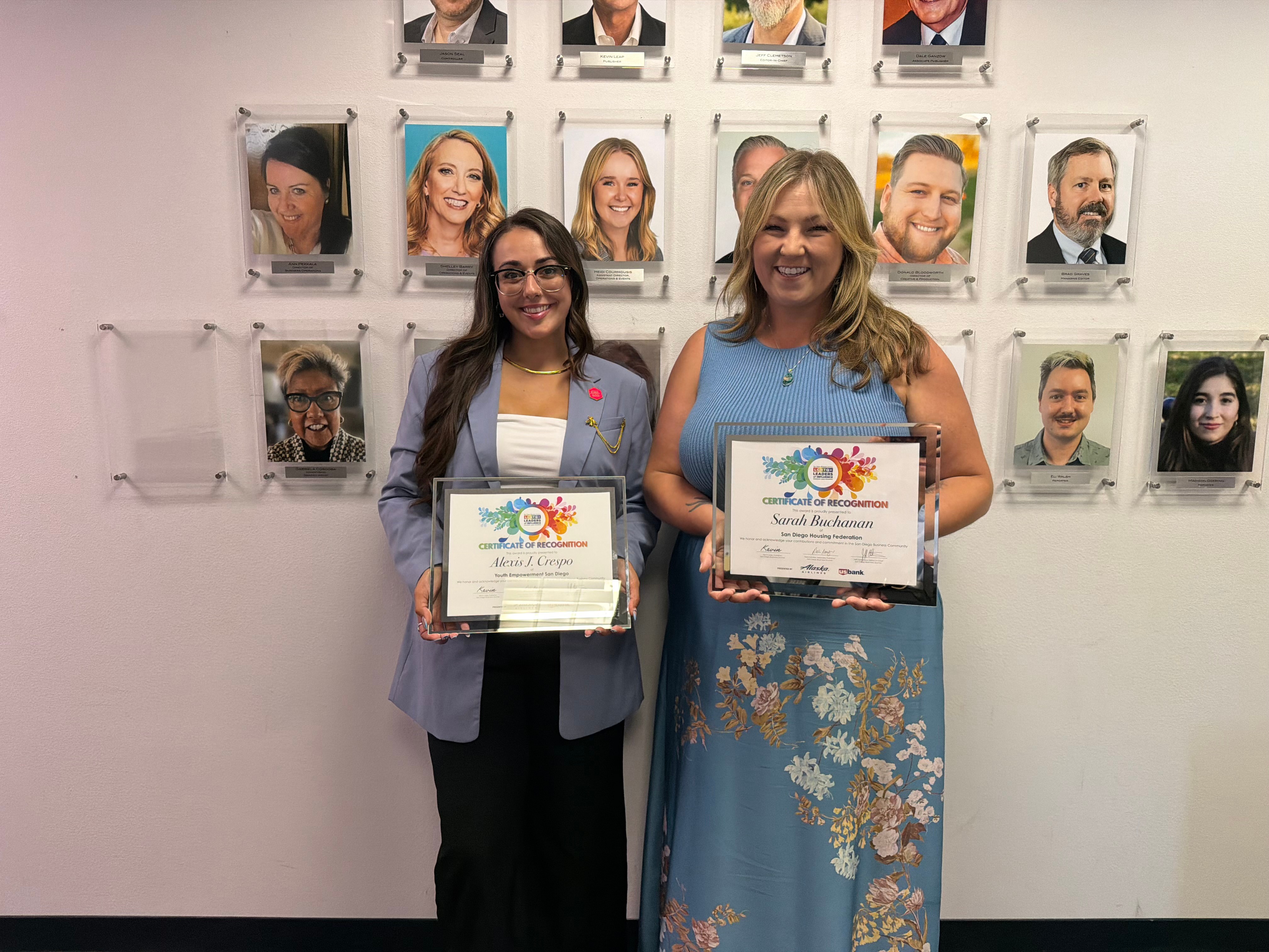 Two women holding certificates, standing in front of a wall with framed photographs of people, smiling.
