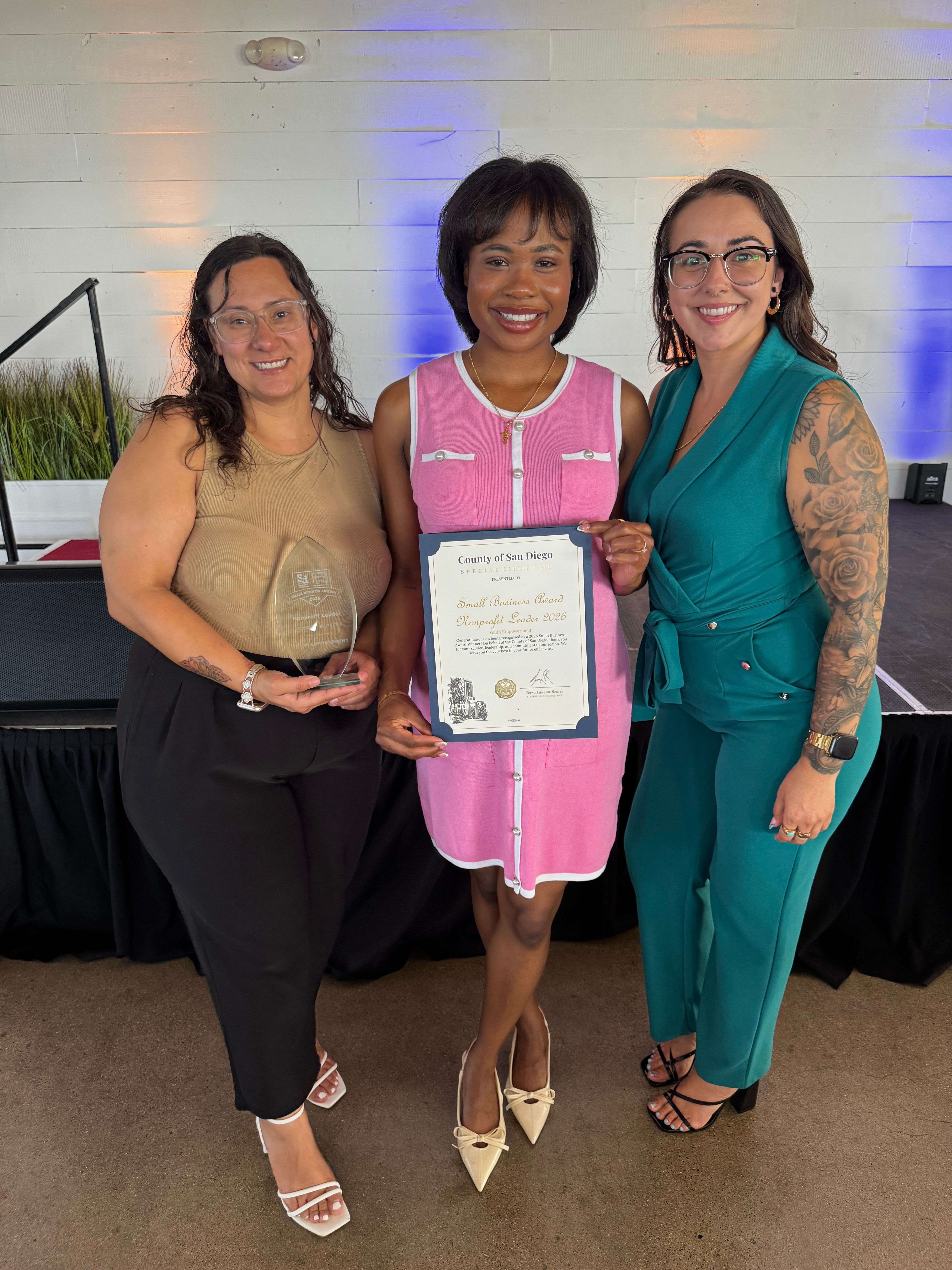 Three women standing together, one holding a certificate, in a room with a plain background and a plant. All are smiling.