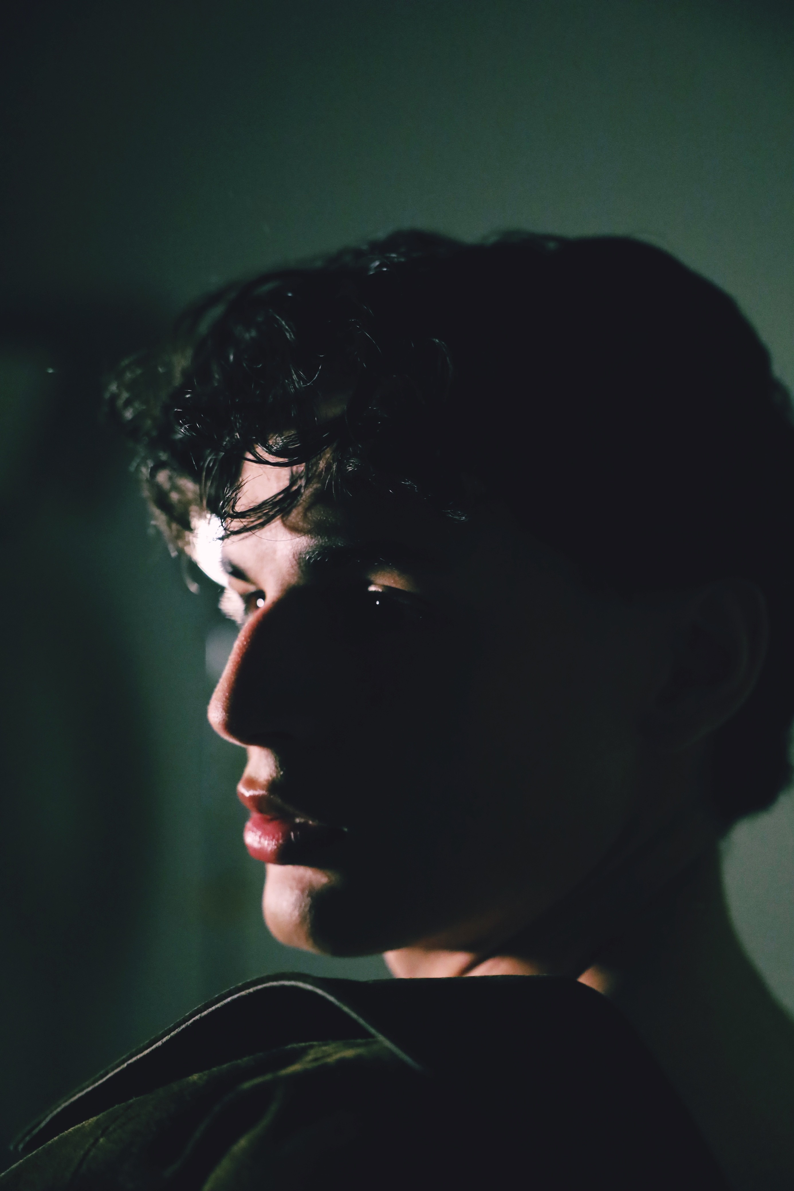 Side profile of a young person with curly hair, dim lighting, and a dark background.