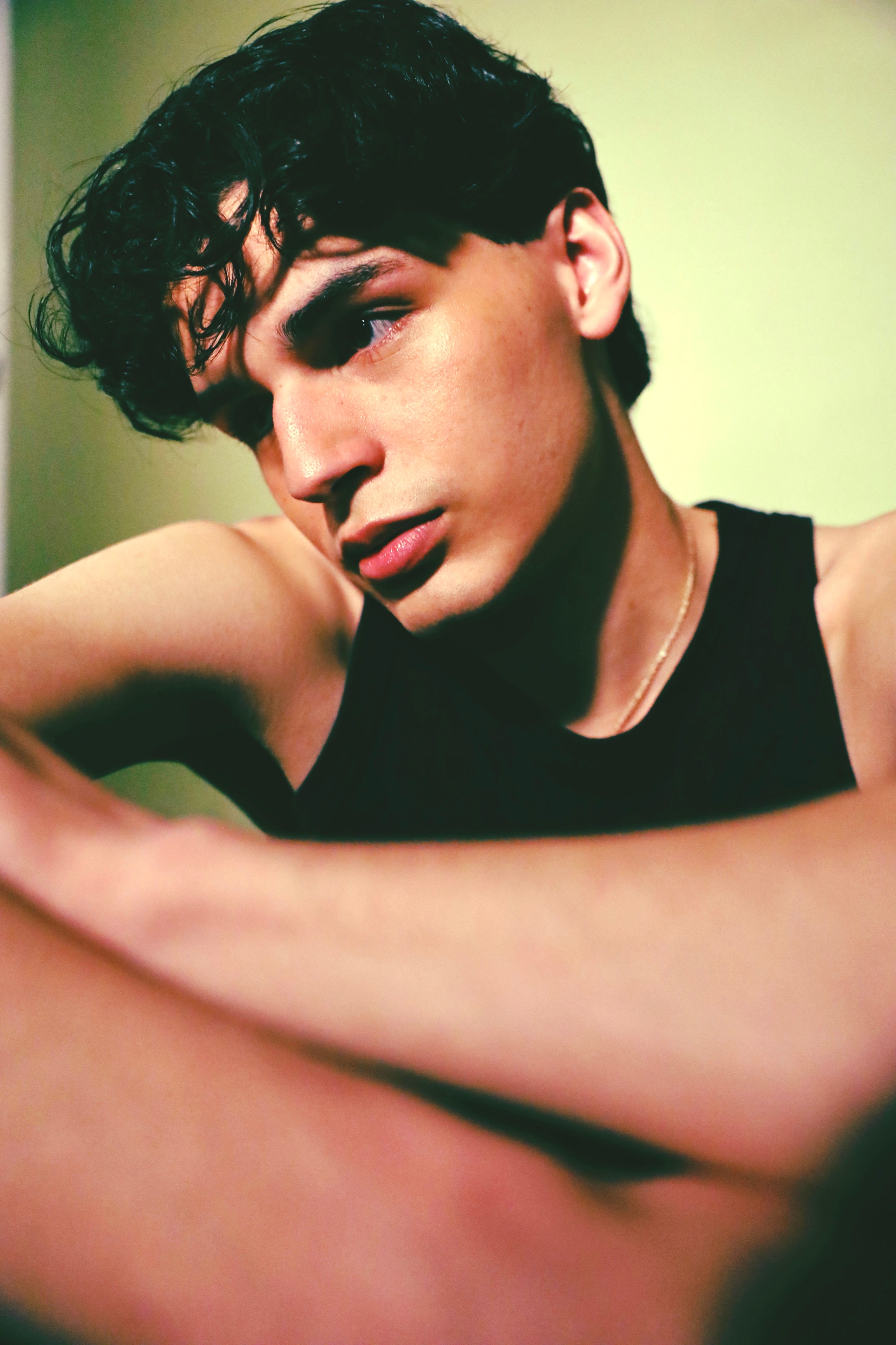 Young person with dark curly hair and sleeveless top, looking at camera, arm resting on surface, neutral background.