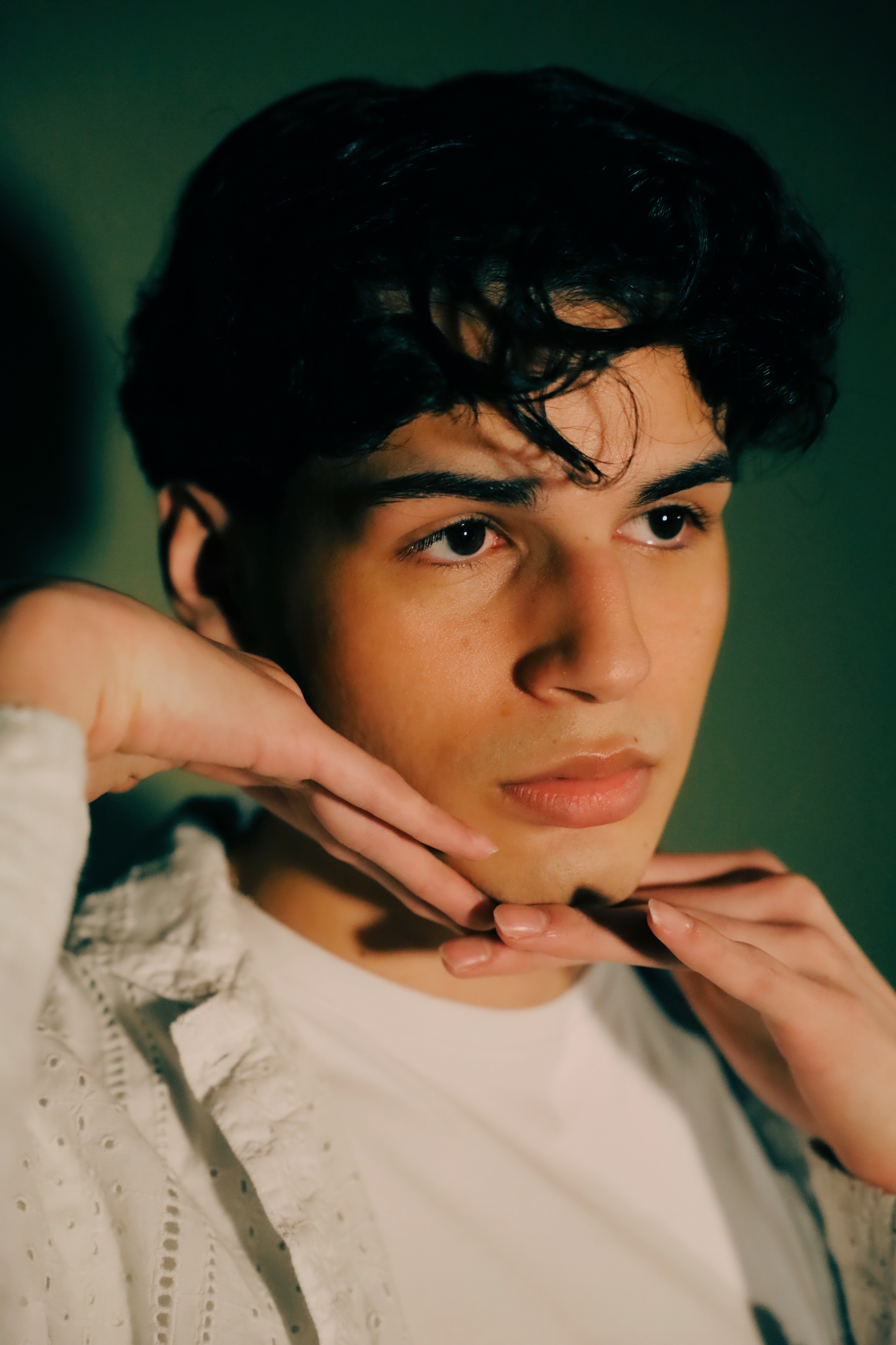 Young person with dark curly hair resting their chin on their hands, looking to the side, against a dark green background.