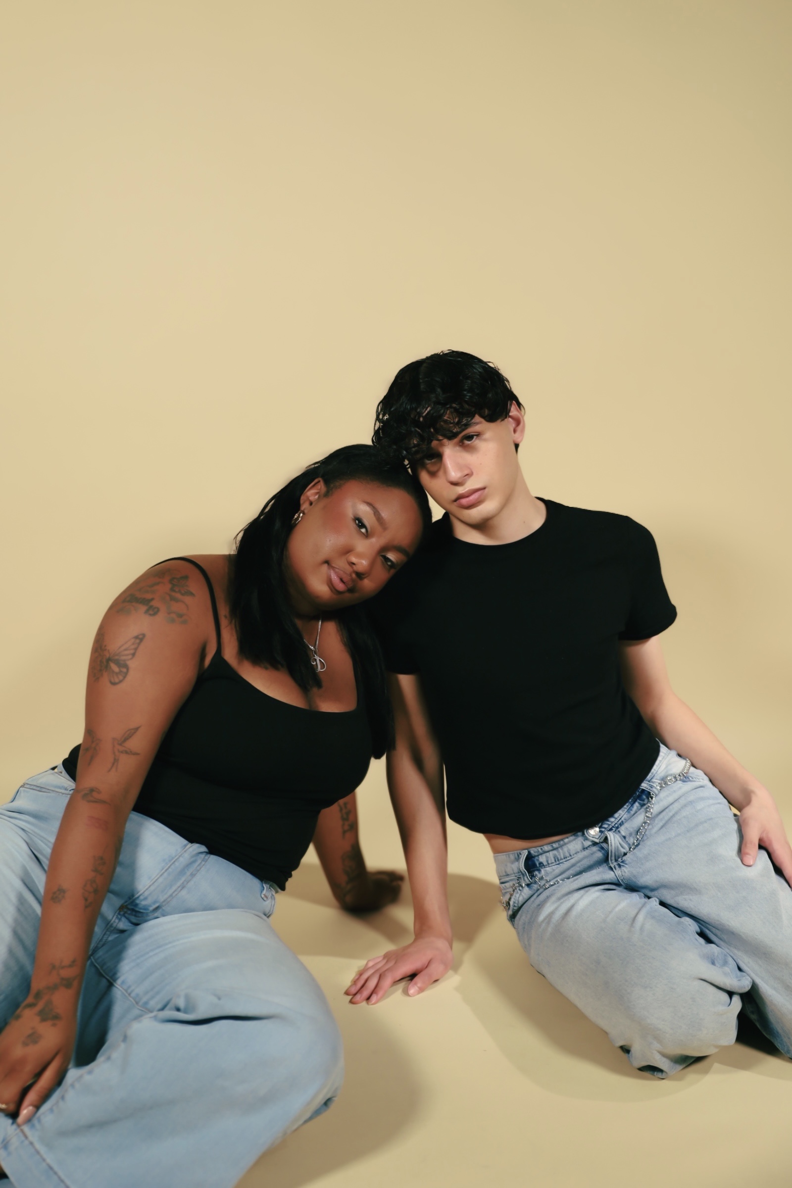 Two people sitting on the floor against a plain beige background, leaning towards each other, wearing black tops and light jeans.