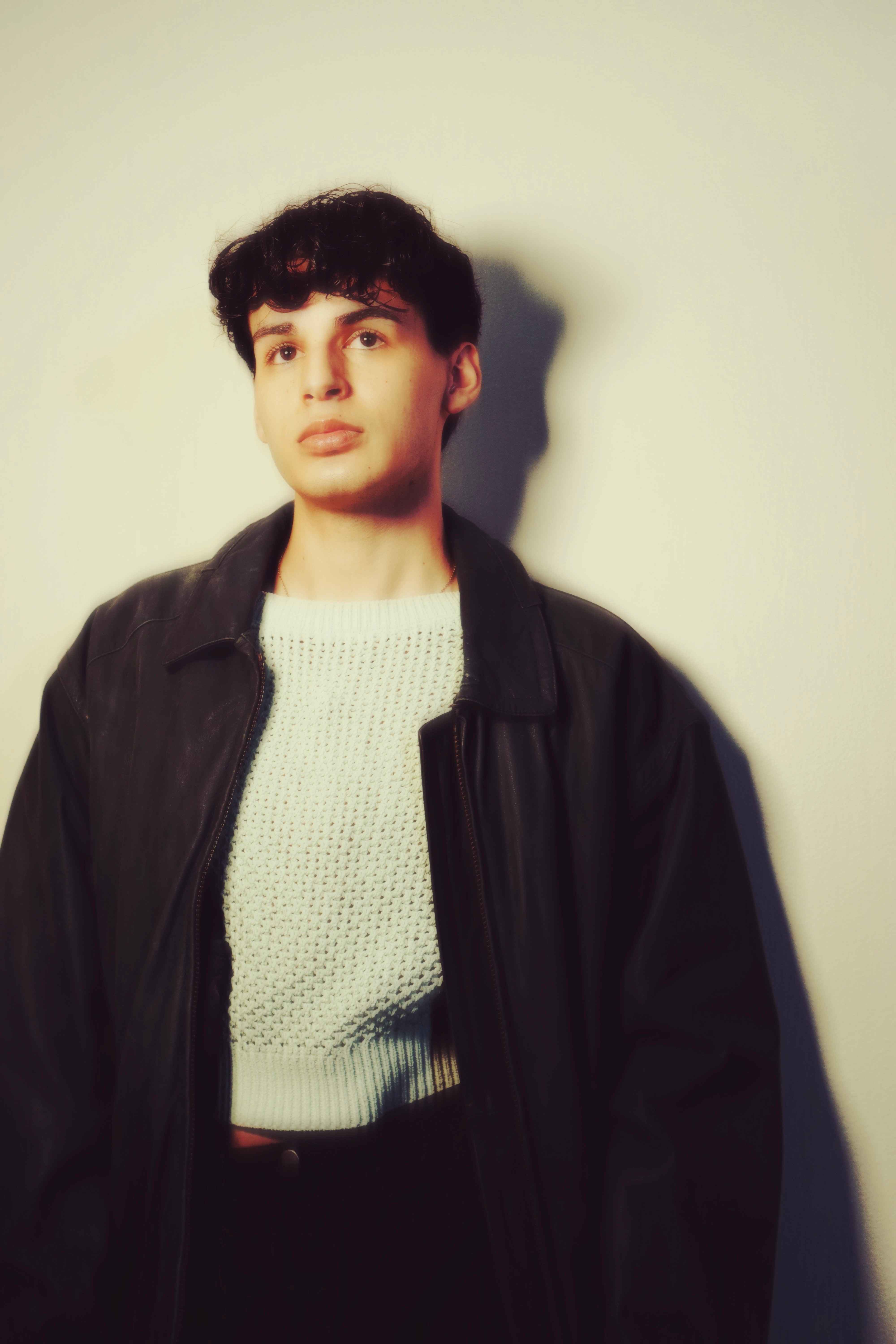 Young man with dark curly hair wearing a black jacket and white sweater, standing against a plain light background.